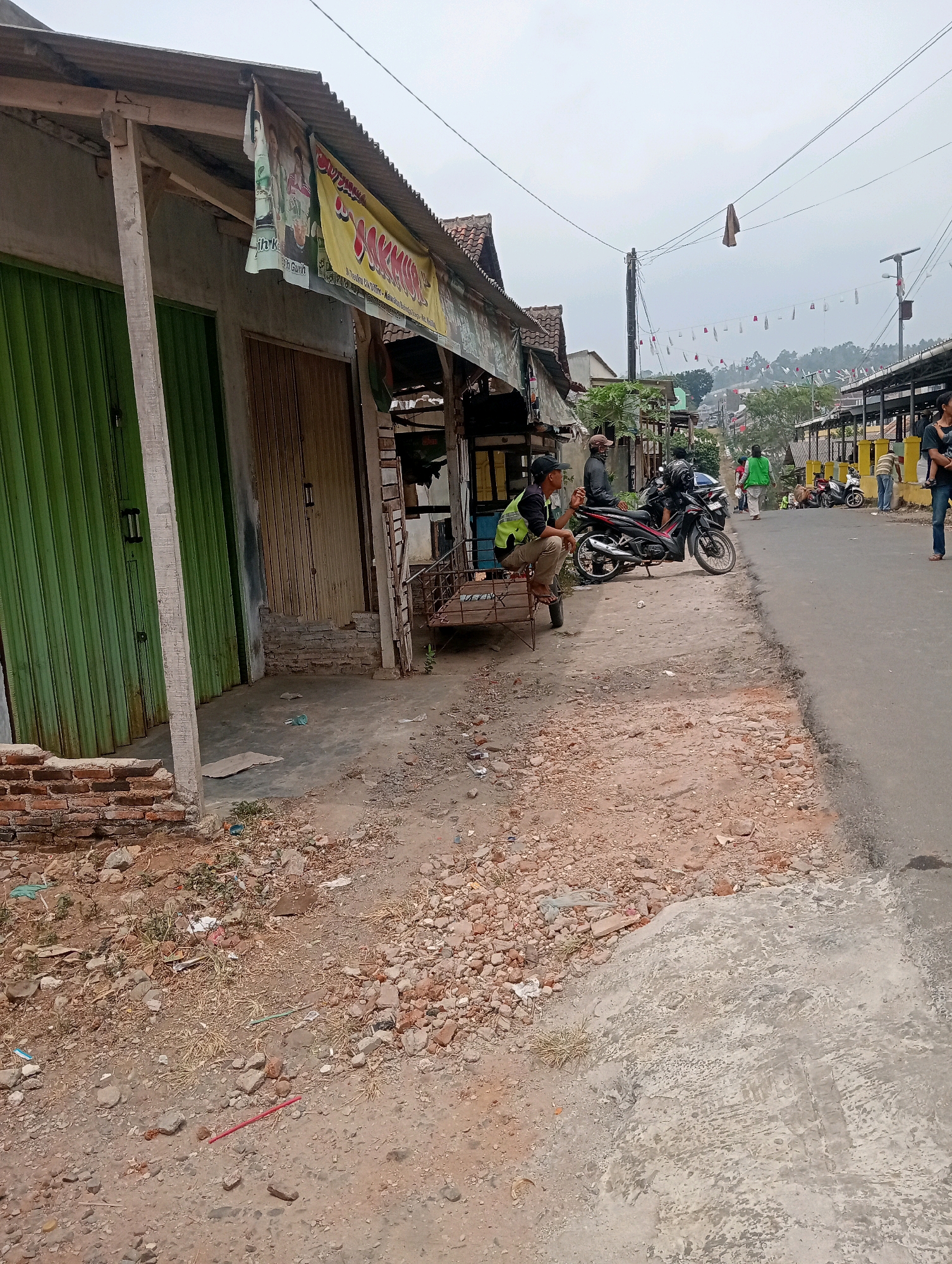 The image depicts a street scene in Bandar Lampung, Indonesia. A dilapidated building with a corrugated metal roof lines the left side of the frame, featuring green and brown roll-up doors. In front of this building, a man wearing a green vest and khaki pants sits on a cart, smoking a cigarette. A motorcycle is parked beside him. The street itself is paved but appears to be in a state of disrepair, with loose gravel and debris scattered along the roadside.
Further down the street, several other motorcycles are visible, along with more buildings and what appear to be stalls or shops. A few other individuals are present in the background, including a woman holding a child. The environment is overcast, and the buildings suggest a somewhat rural or underdeveloped urban area. The overall impression is one of a busy, everyday street in a moderately populated area.