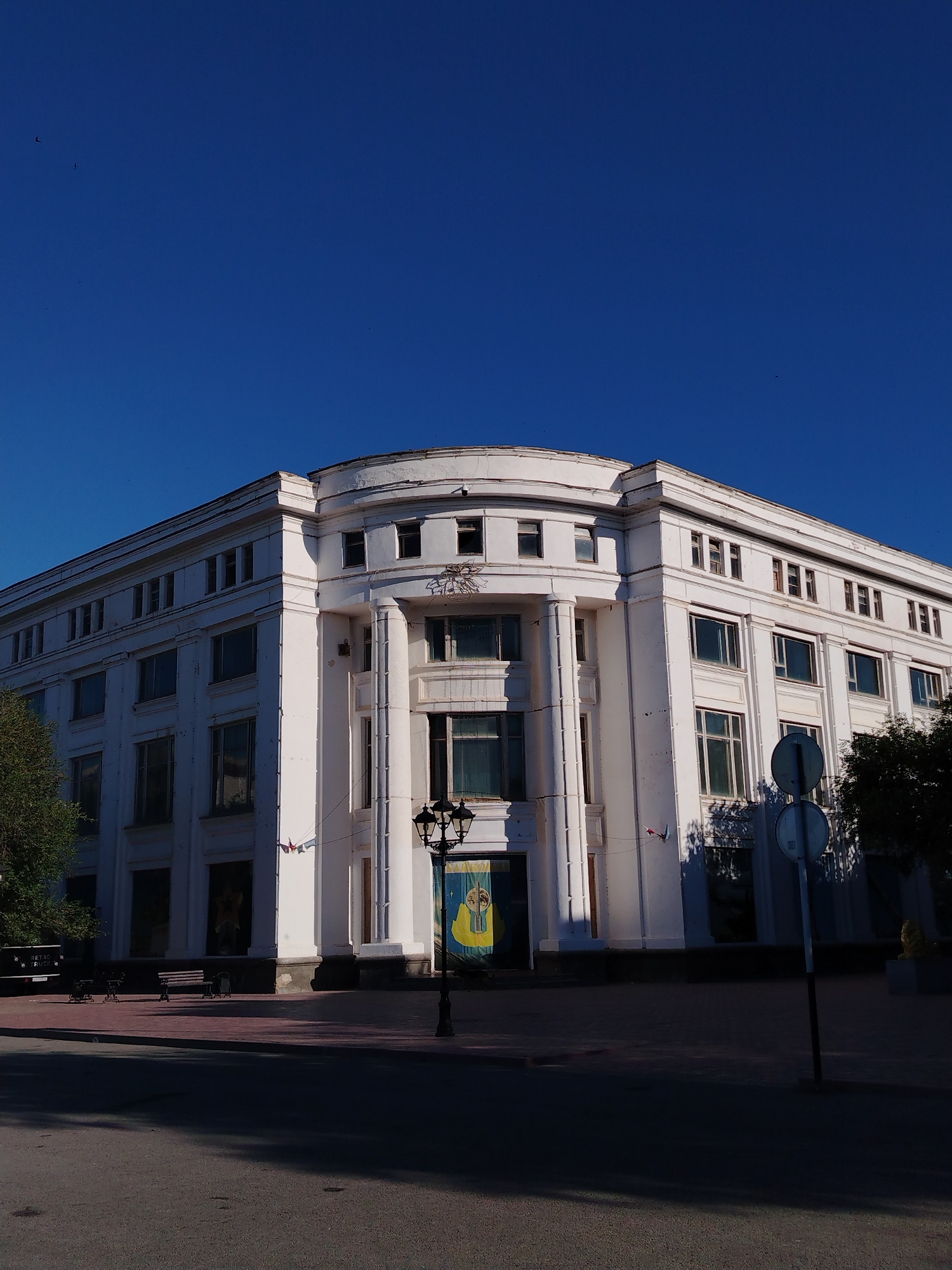 The image captures a low-angle view of a large, white, multi-story building with a curved facade. The building has a neoclassical architectural style, evident from the columns and symmetrical window placement. The sky above is a clear, deep blue with a few small, indistinct dark specks that could be birds or debris. The building's facade is painted white, with visible signs of aging and some peeling paint. Many windows are dark or reflect the sky, suggesting they are either empty or the interior is dimly lit. A tall lamppost with a decorative top stands in front of the building. To the left of the building, a tree with dark green foliage is partially visible. In the foreground, a paved area or sidewalk is present, with benches and what appears to be a traffic sign. The ground in the very front is a shadowed asphalt surface. On the building's facade, near the entrance, a yellow banner with blue and white symbols is displayed. The scene is set on a clear day, with strong sunlight casting distinct shadows. The location is identified as the Palace of Culture in Baikonur, Kazakhstan, a city known for its cosmodrome.