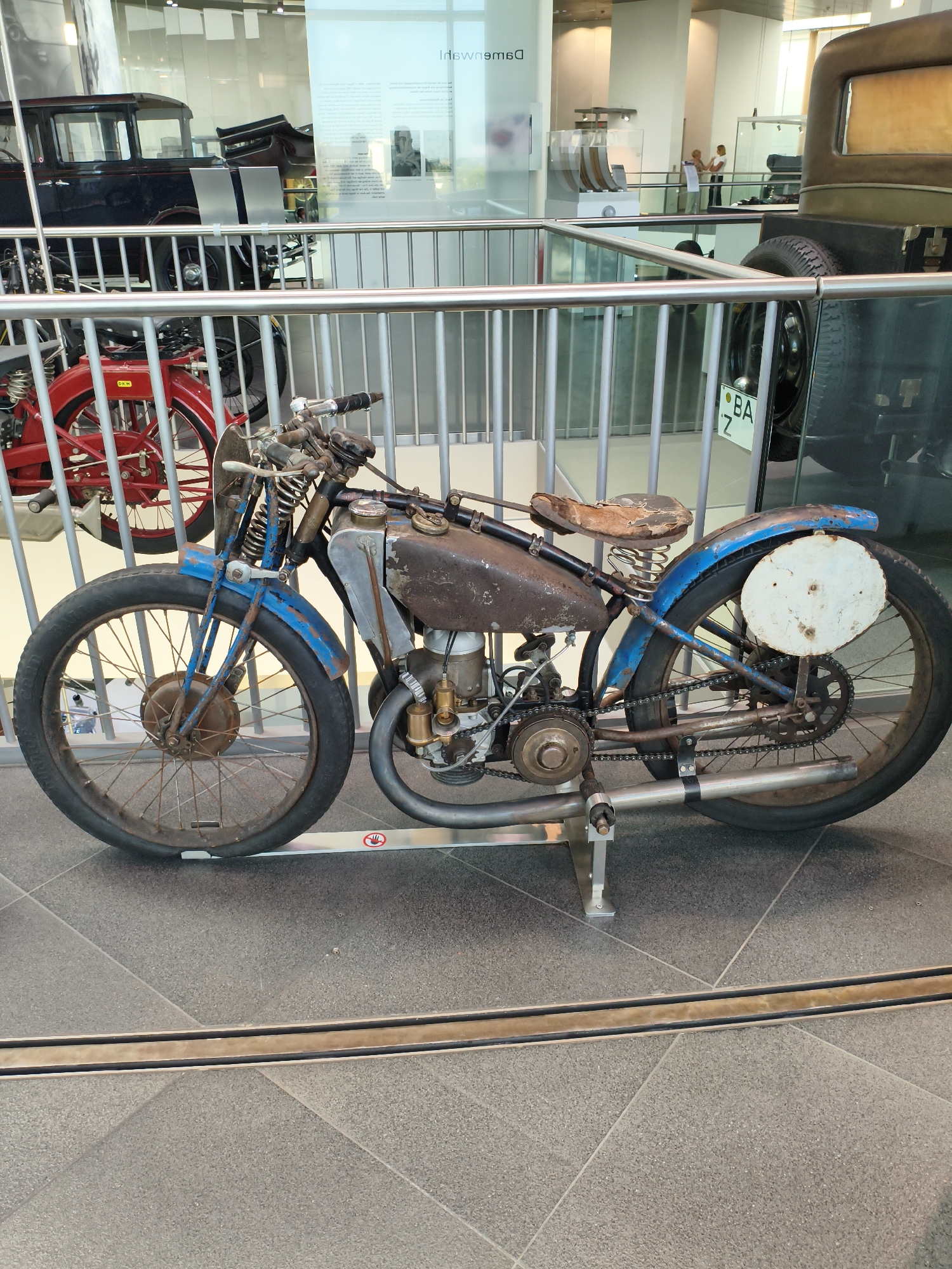 This image depicts a vintage blue and rusty motorcycle displayed in what appears to be a museum. The motorcycle is the central focus, with its weathered frame, worn leather seat, and exposed engine components. It is positioned on a tiled floor and is partially obscured by a metal railing.
In the background, other vintage vehicles are visible, including a dark-colored car and a bright red vehicle, also behind railings. Glass partitions and display cases are present, suggesting an exhibition space. A sign with text in German, "inngas," is partially visible, indicating the location as Ingolstadt, Germany. There are no people clearly visible in the frame interacting with the motorcycle. The overall environment suggests a well-lit indoor exhibition hall dedicated to automotive history.