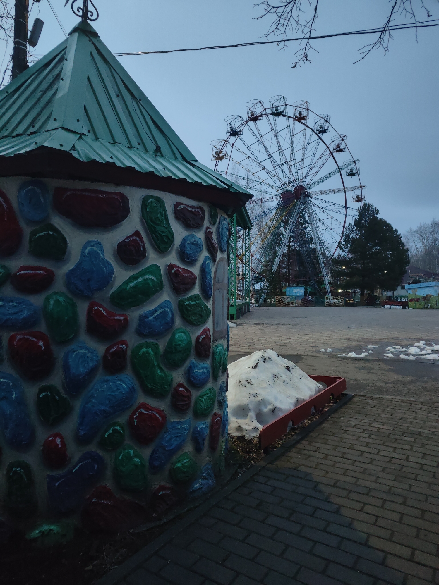 The image captures a section of an amusement park in Chaikovsky, Russia on a cloudy day. A small, stone-textured building with a green, peaked roof sits in the foreground. The building is decorated with painted stones of red, blue, and green. To the right of the building, a pile of snow rests in a red-bordered planter. Beyond the planter is a paved brick walkway. In the background, a large Ferris wheel dominates the scene. Other park structures, trees, and hints of buildings are visible around the Ferris wheel. The sky is overcast.