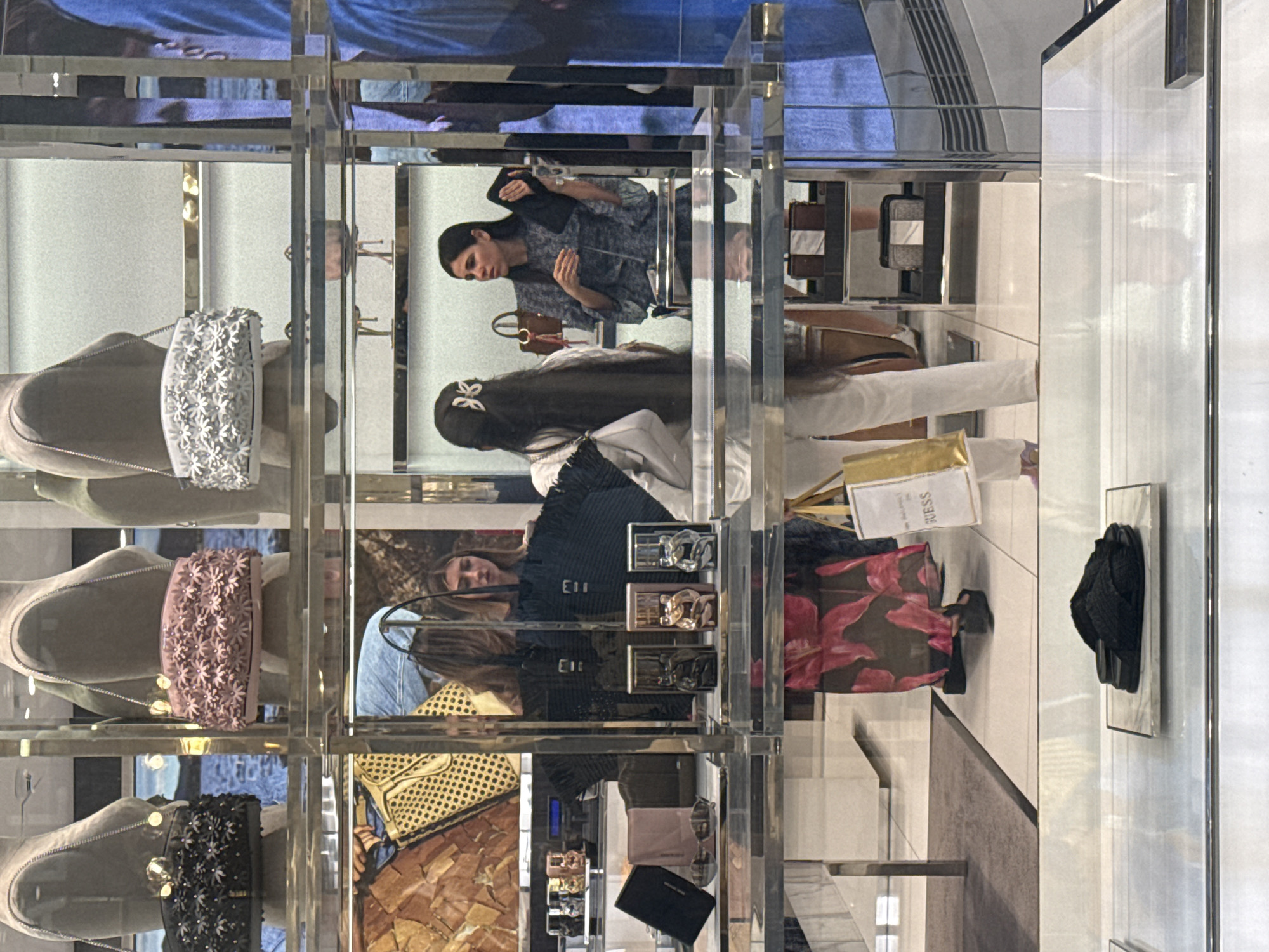 A mirrored display cabinet in Milan, Italy contains handbags. Three women are reflected. One woman holds a black object and wears a gray-blue patterned shirt. A woman with long hair wears a white shirt. A woman with dark hair wears a dark blouse with tassels.