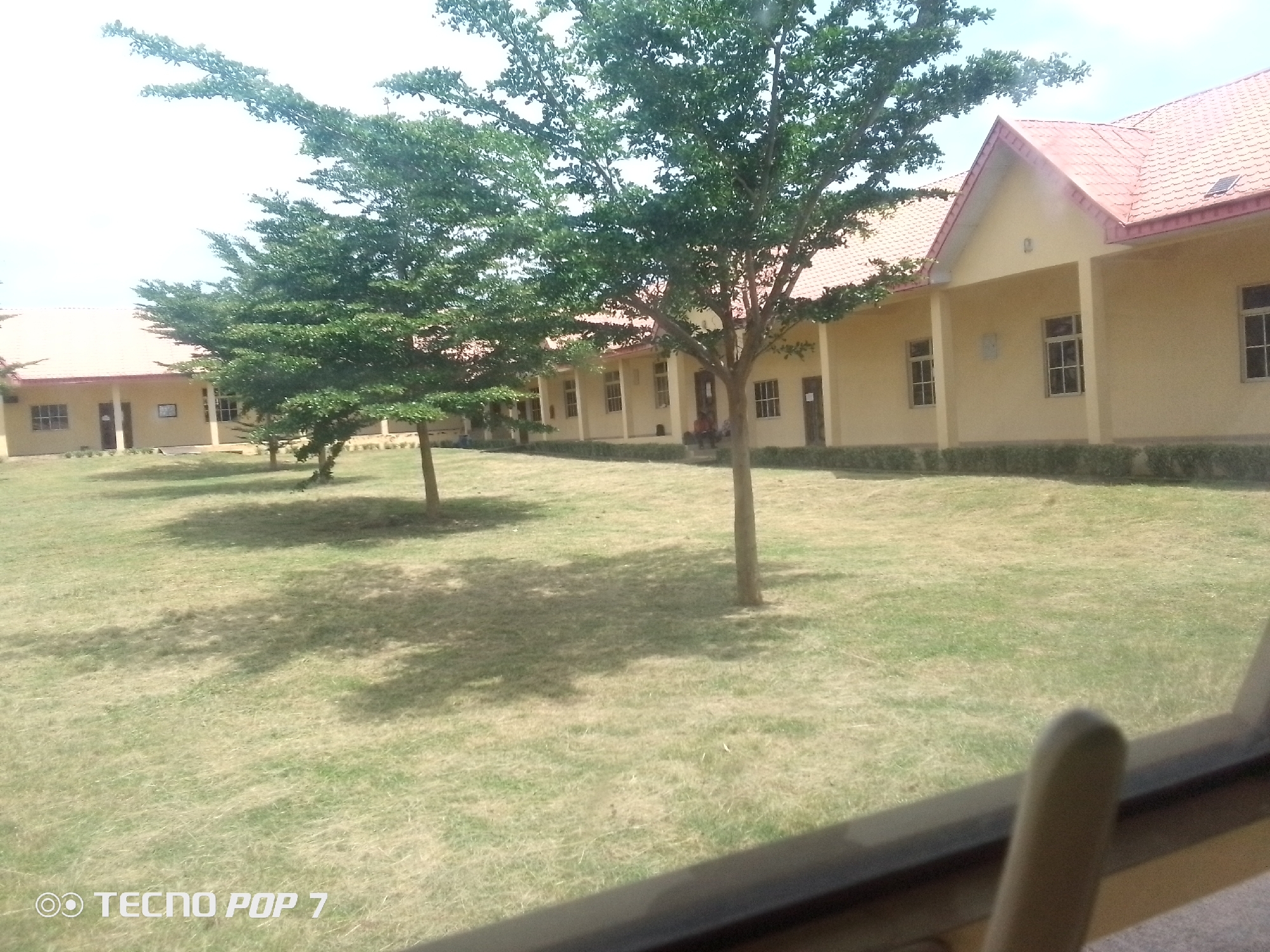 A medium shot captures a row of cream-colored buildings with red tiled roofs, lined by a row of trees with dense green foliage. The buildings have several windows and doors, and a shaded porch area. A patch of green grass covers the foreground, with scattered trees casting shadows. A watermarked "TECNO POP 7" is visible in the bottom left corner. The scene appears to be located in Eleweran, Nigeria, likely on the grounds of an institution or complex. No individuals are clearly visible or engaged in any notable actions.