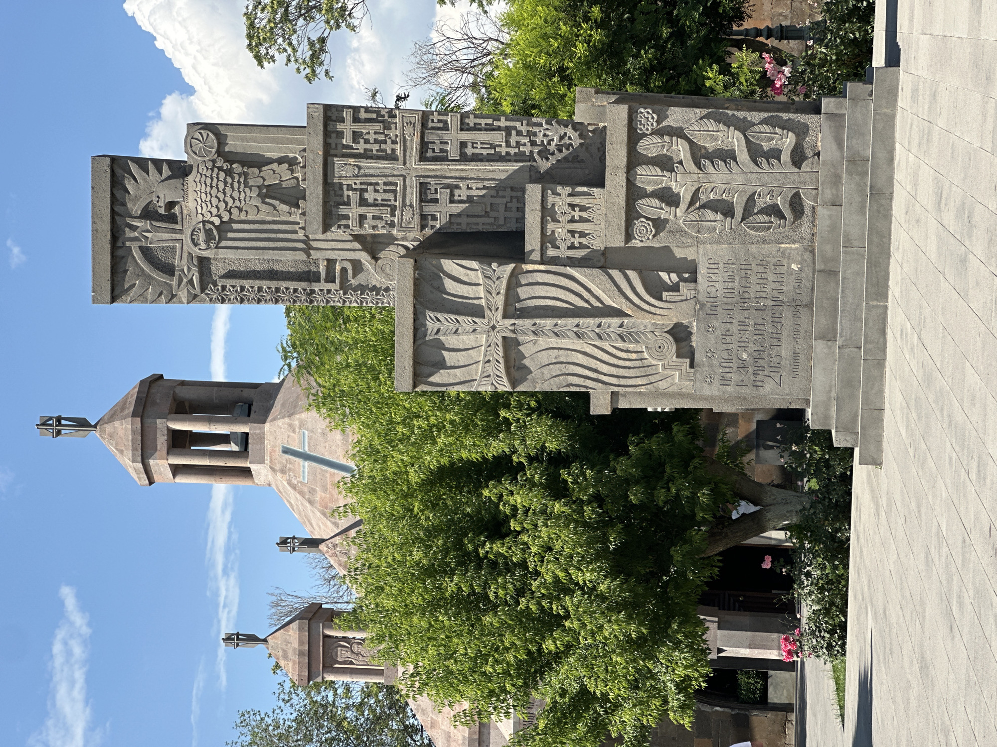 The image shows a memorial monument in front of the Mother See of Holy Etchmiadzin in Vagharshapat, Armenia. The monument, made of carved stone, features intricate designs including crosses, an eagle, and what appears to be the Tree of Life. In the background, the distinctive architecture of the Mother See is visible, with its stone domes and bell towers topped with crosses. Lush green trees and flowering bushes surround the monument and the church. The sky is blue with scattered white clouds. The scene is bathed in sunlight, suggesting it is daytime. No people are clearly visible in the immediate vicinity of the monument, though there are hints of individuals near the entrance to the church. The ground is paved with light-colored stone tiles.