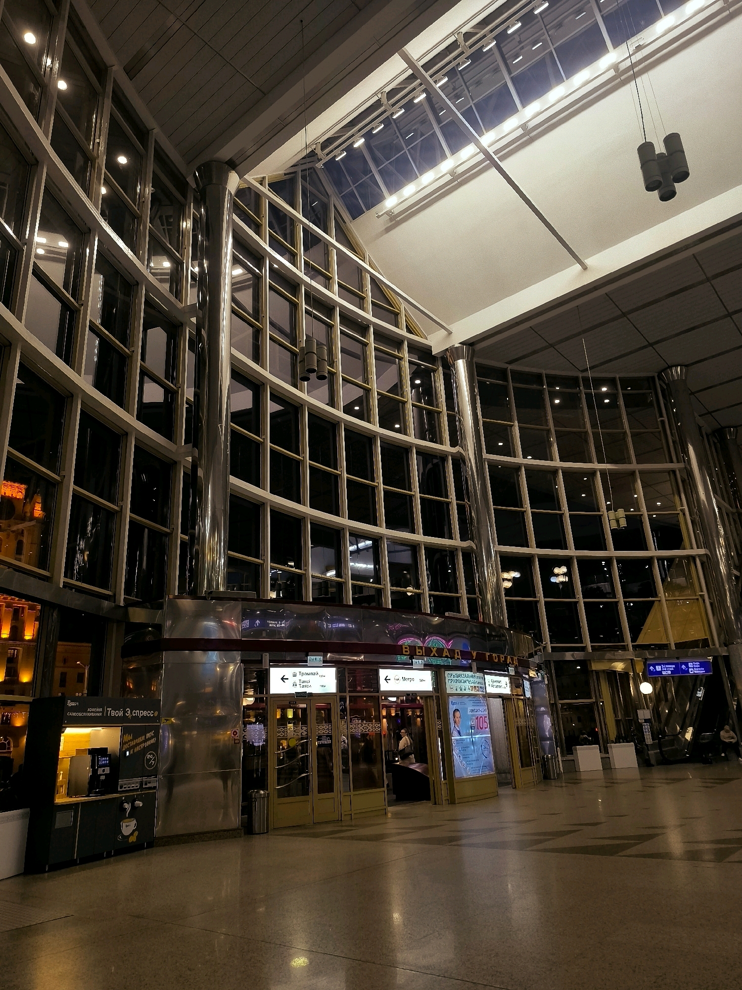 The image depicts the interior of a modern building, likely a transportation hub like a train station or airport terminal, in Minsk, Belarus. The architecture features a grand, multi-story atrium with extensive glass paneling and metallic support columns. Sunlight streams in through the large windows and the transparent ceiling, illuminating the polished floor and casting reflections. In the foreground, there are several kiosks, one of which appears to be a coffee stand with a prominent sign in Cyrillic. Further into the space, glass-enclosed entrances lead to different areas, marked with directional signs also in Cyrillic. A few individuals are visible, some walking and others seated in waiting areas. The overall ambiance is one of contemporary design and functional public space.