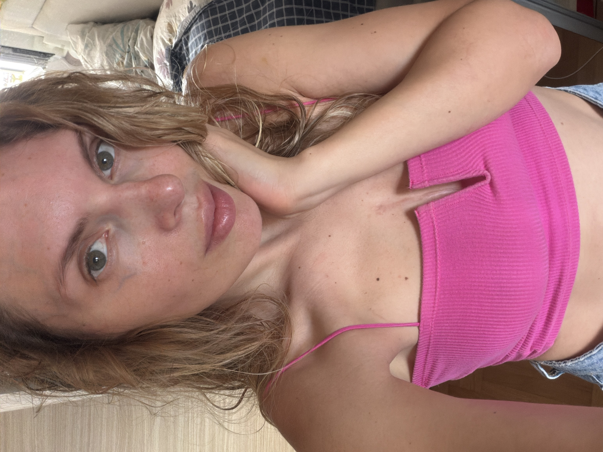 A young woman with long, wavy blonde hair and green eyes is depicted in a close-up, tilted selfie. She is wearing a bright pink ribbed crop top with thin shoulder straps, and a small portion of light blue denim shorts is visible on her right side. Her left arm is bent, with her hand resting near her neck/shoulder, contributing to the casual, self-taken nature of the photograph.
Her gaze is directed towards the camera with a neutral to slightly contemplative expression, and her lips appear glossy. Freckles are visible on her face and shoulders, and a small scar or mark is present on her sternum area.
The background suggests an indoor setting, likely a bedroom or a relaxed living space, with pillows and bedding partially visible behind her head. The lighting appears to be natural daylight. There is no visible text in the image. The setting is in Budva, Montenegro.