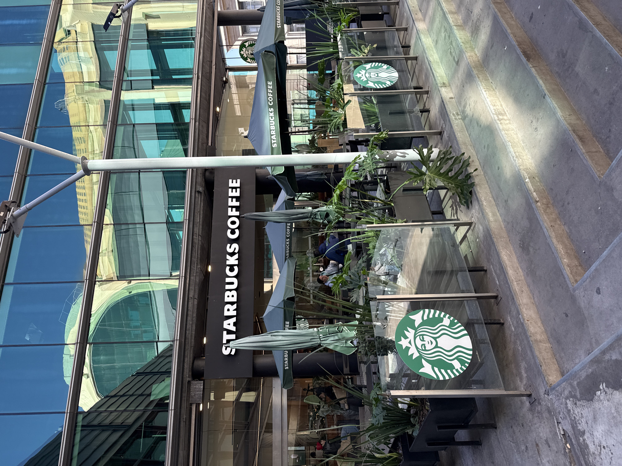 The image displays an outdoor scene of a Starbucks coffee shop, set within an urban environment, identified as being in Buenos Aires, Argentina. The coffee shop occupies the ground level of a modern building characterized by large, reflective glass windows that mirror the surrounding architecture.
In the mid-ground, an outdoor seating area is furnished with tables and chairs, shaded by several green Starbucks umbrellas. Various potted plants and lush greenery are interspersed throughout the patio, enhancing the outdoor ambiance. A few people are visible seated at the tables, seemingly patrons enjoying beverages or conversation.
A large, vertical sign on the building clearly displays "STARBUCKS COFFEE" in white lettering against a dark background. The iconic circular Starbucks siren logo is prominently featured on the green umbrellas and on transparent glass partitions that enclose the seating area.
To the right of the coffee shop's patio, a series of wide, concrete steps or tiered platforms ascend diagonally upwards, suggesting a multi-level public space. The reflective glass facade of the building captures detailed reflections of nearby structures, including an older, light-colored building with a distinct domed or rounded architectural element, showcasing a blend of architectural styles in the city. The reflections also indicate a clear, blue sky.
The scene is brightly illuminated, suggesting it is daytime with sunny weather. The overall impression is that of a lively urban coffee shop setting.