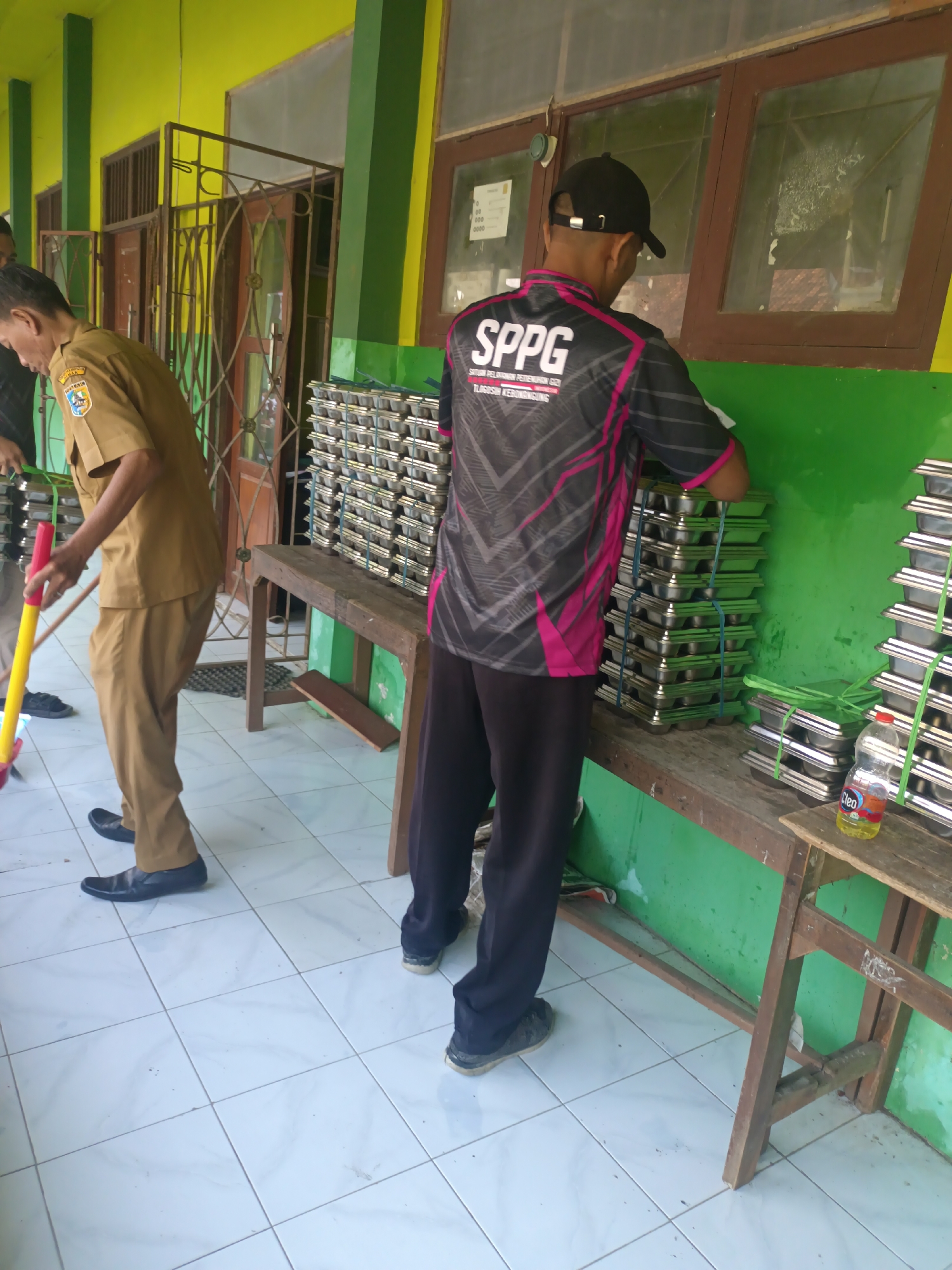 In the city of Sarimulyo, Indonesia, two individuals are engaged in activity near a building with lime green walls and wooden framed windows. The first individual, wearing a tan uniform, is sweeping the tiled floor with a yellow and red broom. To the right of the first individual, a second person, dressed in a black shirt with pink accents and a black baseball cap, is organizing stacks of metal trays. The trays are neatly piled and secured with green and blue ties. One stack of trays is on a wooden bench, and another is on a rustic wooden table. A partially full plastic bottle of "Cleo" brand water is visible on the table. The environment appears to be an outdoor or semi-outdoor space, possibly a school or community center, given the building's architecture and the presence of the uniformed individual. The floor is tiled in a light color with a grid pattern.