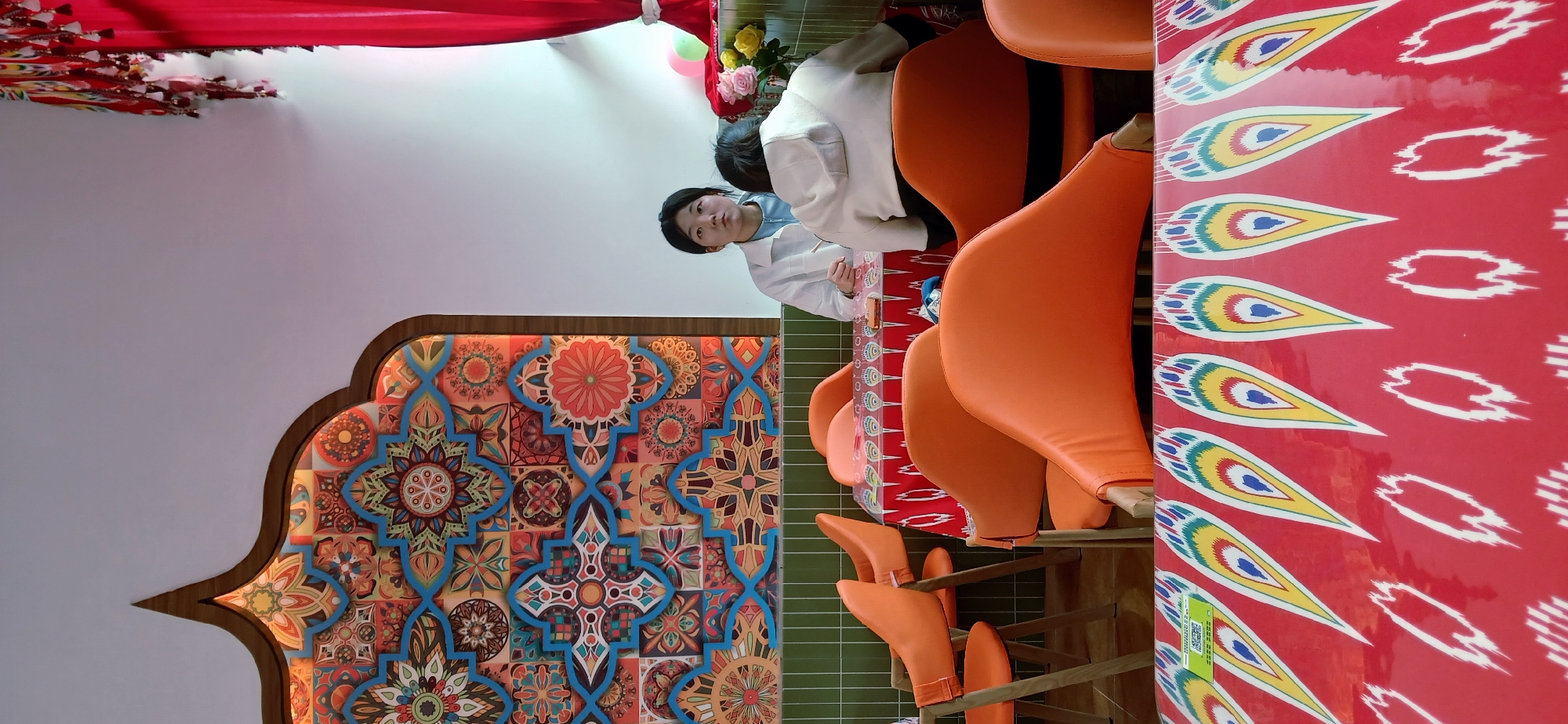 The image depicts the interior of a restaurant in Chongqing High-tech Zone, China. A woman in a white jacket and blue top is seated at a table with a red patterned tablecloth. She is looking directly at the camera. There are orange chairs around the table. To the left, there is a wall decoration with a floral and geometric pattern. Red curtains are partially visible at the top of the image. The restaurant has a patterned wall and patterned tablecloths.
