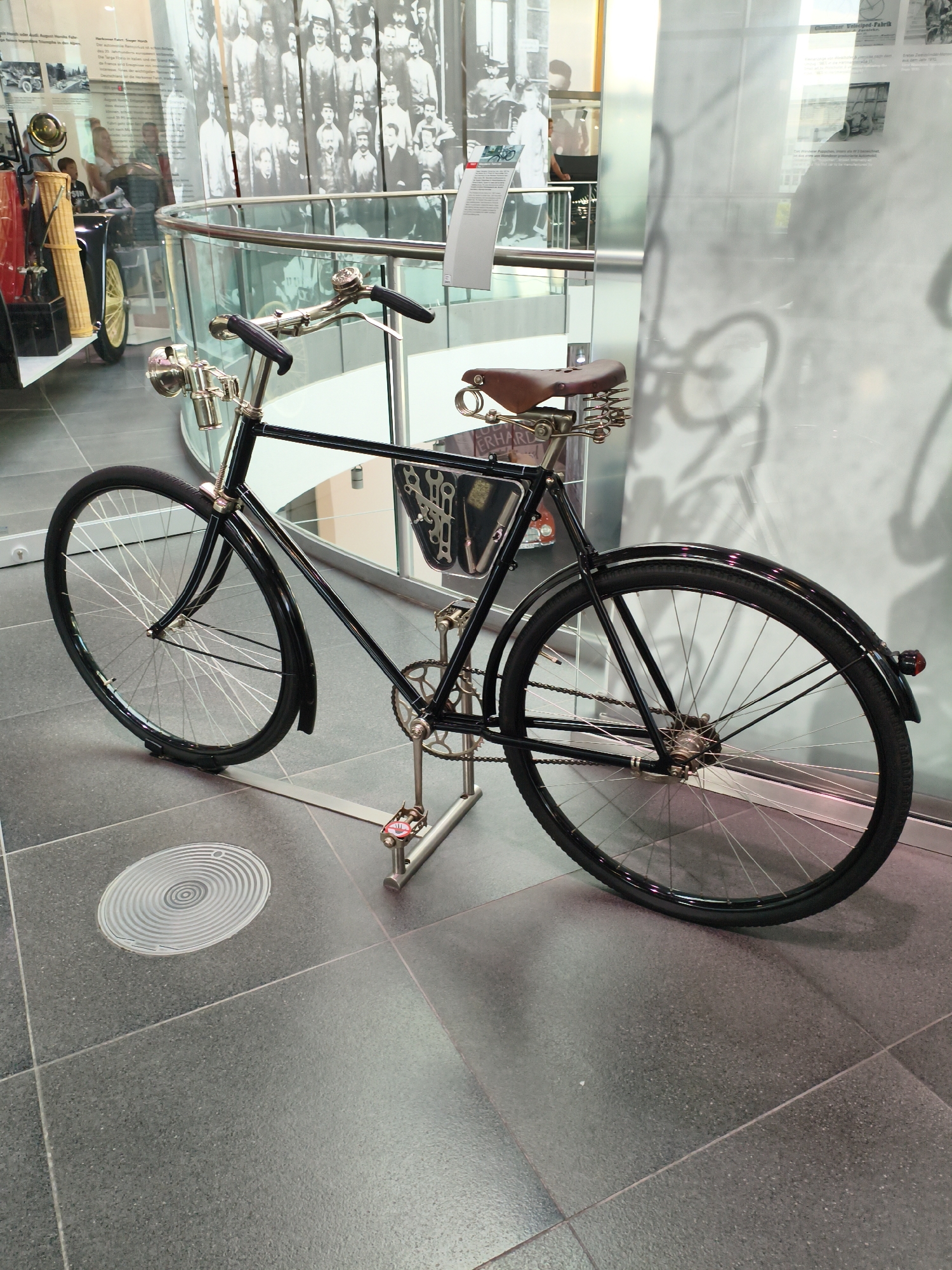This image depicts a vintage black bicycle on display in a museum. The bicycle is positioned on a metal stand and features a brown leather saddle, chrome handlebars with black grips, and a visible chain and gears. The background shows a glass enclosure with historical photographs and informational text. The floor is tiled in a dark, speckled pattern. The context of Ingolstadt, Germany, suggests this may be the Audi Museum mobile, given the presence of historical vehicles and exhibits. No people are clearly visible interacting with the bicycle.