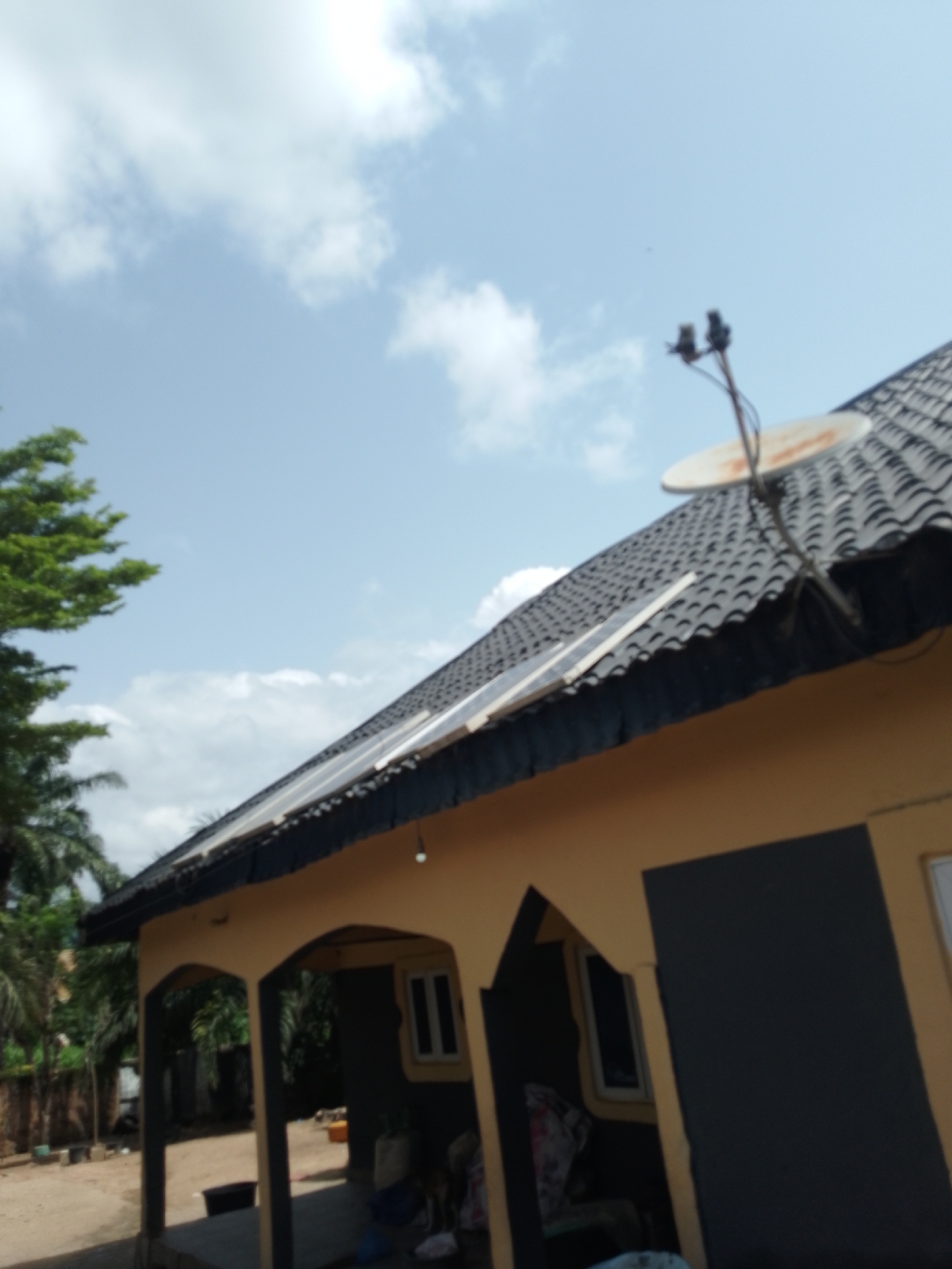 The image shows a close-up, angled view of a residential building with a dark, tiled roof. The building's exterior wall is painted a light yellow. Several solar panels are visible mounted on the roof. A satellite dish with a central LNB is also present on the roof, attached to a pole. To the left of the building, lush green foliage from trees, likely palm trees, is visible against a bright, partly cloudy sky. The foreground shows the front of the house with arched openings leading to an interior space. A small light fixture is attached to the underside of the roof overhang. The environment appears to be a residential area. There are no people visible in the image. The image quality is somewhat blurry, suggesting it was taken with a low-resolution camera or in low light conditions. The scene is consistent with a dwelling in Serti, Nigeria, given the climate and architectural style.