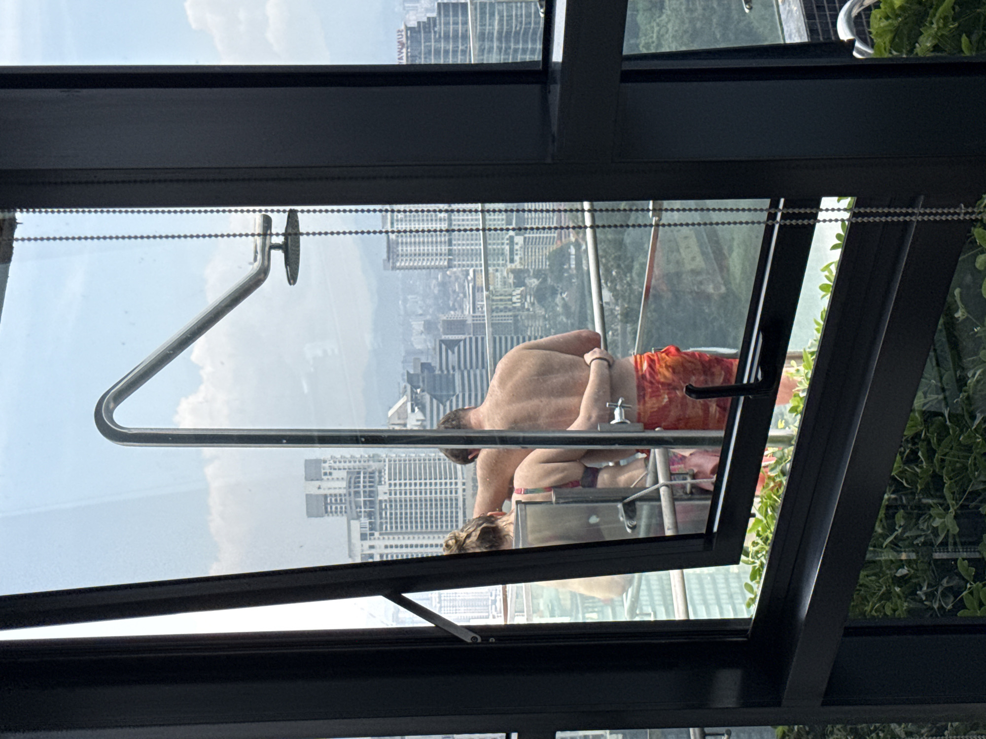 The image shows a window in Kuala Lumpur, Malaysia, looking out at the city skyline. A couple is visible through the window; the man is shirtless and wearing orange swimming trunks, and the woman has blonde hair in a bun. They are sitting on a deck chair in what appears to be an outdoor area with a glass railing. The window frame is black and partially open, with a window handle visible in the top left corner. The exterior surroundings include city buildings, greenery, and an overcast sky.