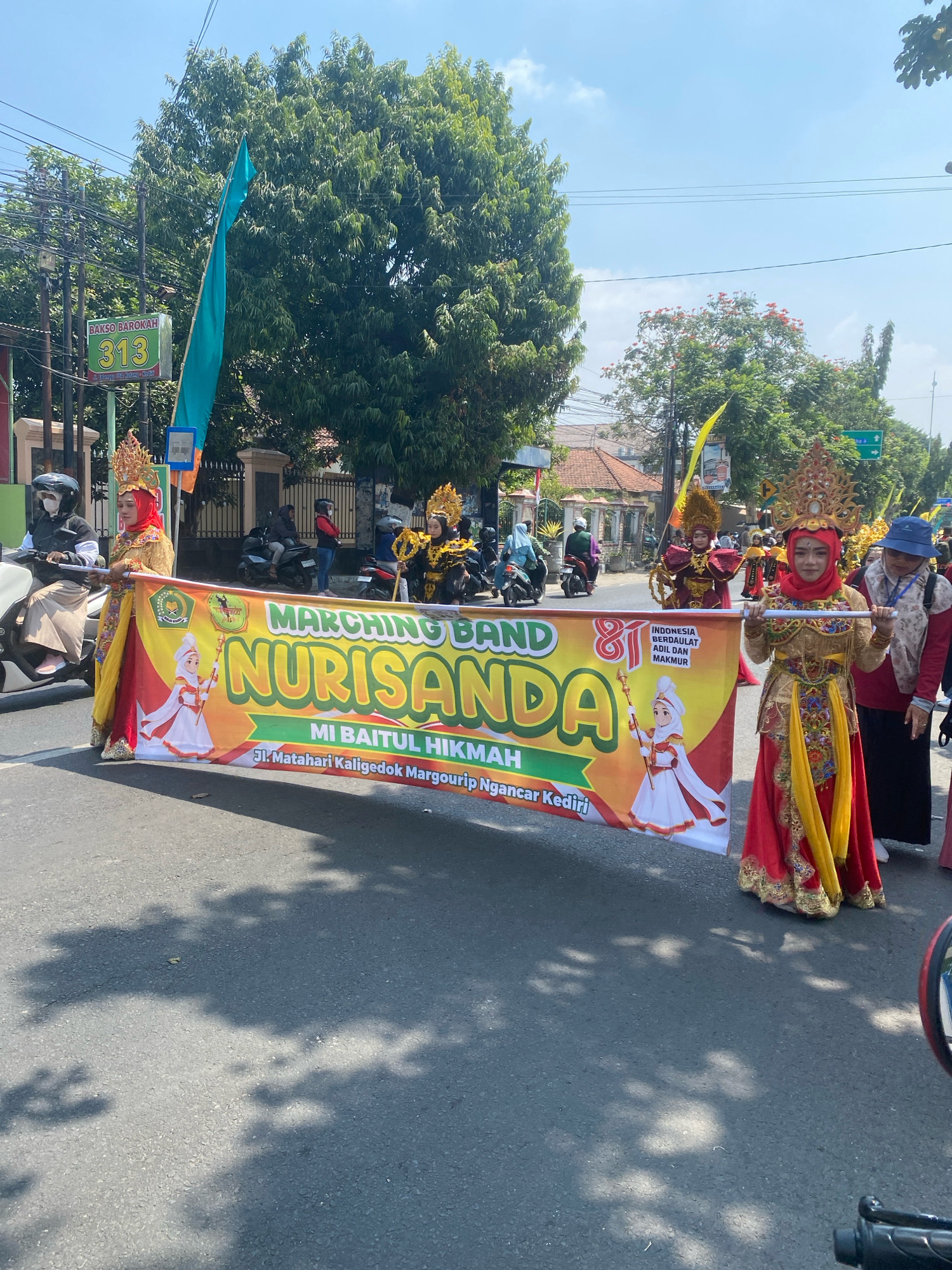 A daytime, eye-level, medium shot captures a lively street parade in Ngasem, Indonesia. A large banner, held by two individuals in ornate, traditional attire, proclaims "MARCHING BAND NURISANDA MI BAITUL HIKMAH JL. Matahari Kaligedok Margourip Ngancar Kediri". The banner features a cartoon illustration of a young girl in a white and red dress holding a staff. The woman on the right wears a red hijab and a gold and red ensemble with elaborate headwear. The woman on the left, partially obscured by the banner, also wears a red hijab and a similar outfit. Behind them, other participants in colorful costumes, some with golden headpieces, are visible. A person on a scooter, wearing a white hijab and a black jacket, is in the foreground on the left. In the background, more people on motorbikes and on foot, along with trees and buildings, line the street. The sky is clear and blue. A green sign with the number "313" and text that reads "BAKSO BAROKAH" is also visible on the left side of the image, along with a turquoise flag and utility poles.