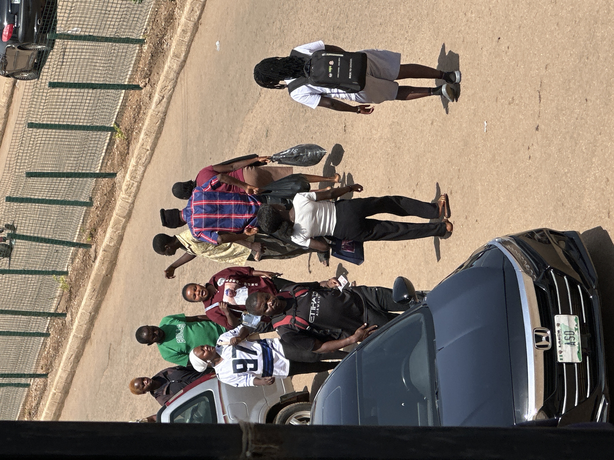 This media file depicts a group of people gathered on a paved surface, likely a street or parking lot, in Bukuru, Nigeria. The image is shot from an elevated perspective, looking down on the scene. The subjects are a mix of men and women, some walking and some standing, with varying levels of engagement.
One individual, a young person with dreadlocks, is walking away from the camera, wearing a white t-shirt, grey shorts, and a backpack with the text "BACK TO SCHOOL" visible. Several other individuals are seen walking in a line, carrying bags, some of which are black plastic.
In the foreground, a dark-colored car is parked, with its front grille and license plate visible. The license plate has text that appears to be "ASD" in green letters on a white background, with other smaller text above it. Another vehicle, a silver SUV, is partially visible behind the dark car, with some of the people standing near it.
The setting appears to be outdoors, under daylight, suggesting a daytime scene. The ground is a light brown, paved surface, with some scattered debris. A chain-link fence and some vegetation are visible in the background. The overall mood seems neutral, with no strong indications of specific emotions. The activity appears to be people moving about, possibly commuting or congregating.