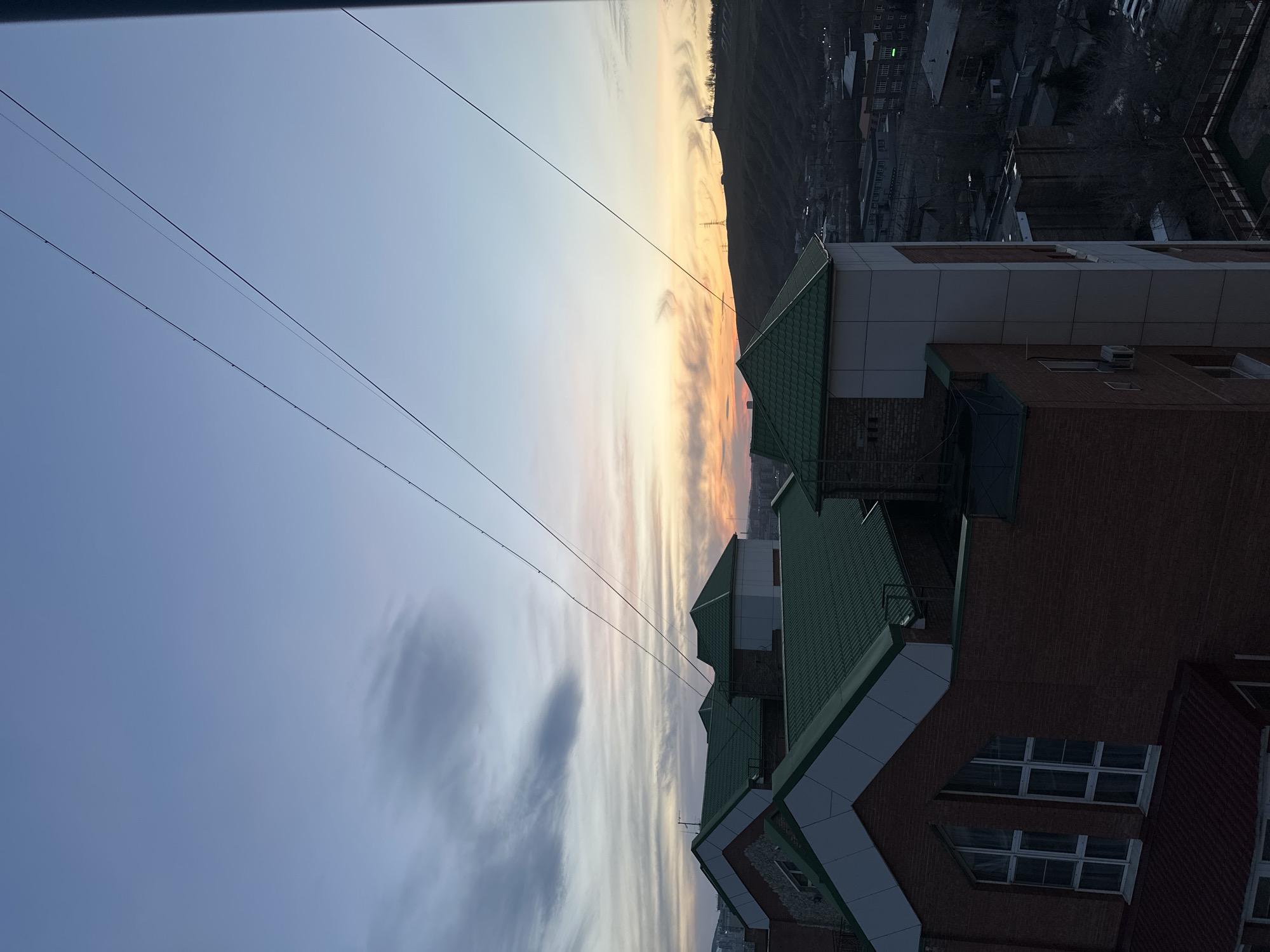 A high-angle view captures a cityscape and sky during either sunrise or sunset over Krasnoyarsk, Russia. The image is oriented vertically (rotated 90 degrees clockwise from typical landscape), presenting a horizon that runs diagonally from the lower left to the upper right.
In the upper portion of the frame, spanning the diagonal horizon, the sky displays a dramatic gradient. On the left, it transitions from a deep, somewhat hazy blue-grey to softer pale blues and whites, with thin, streaky clouds. Towards the right, closer to the horizon line, the sky glows with warm oranges, yellows, and pinks, characteristic of golden hour light.
Running across the mid-upper section of the image, against the sky, are several thin, dark power lines or communication wires, creating parallel horizontal lines.
In the lower and right parts of the frame, parts of multi-story buildings are visible. These structures feature distinctive dark green, pitched roofs and reddish-brown brick facades, often accented with white or light grey siding. Some windows are discernible on the brick walls. The architectural style suggests a modern or recently renovated appearance.
Beyond these immediate buildings, in the distance, a dark, undulating silhouette of hills or mountains stretches along the horizon, contrasting sharply with the bright sky. Below the hills, a sprawling urban or suburban landscape is dimly visible, fading into twilight or pre-dawn darkness. Small, faint lights might be visible within this distant urban sprawl. No specific activity or human subjects are depicted in the scene. There is no visible text. The overall atmosphere is serene and tranquil, capturing the fleeting beauty of daybreak or dusk.