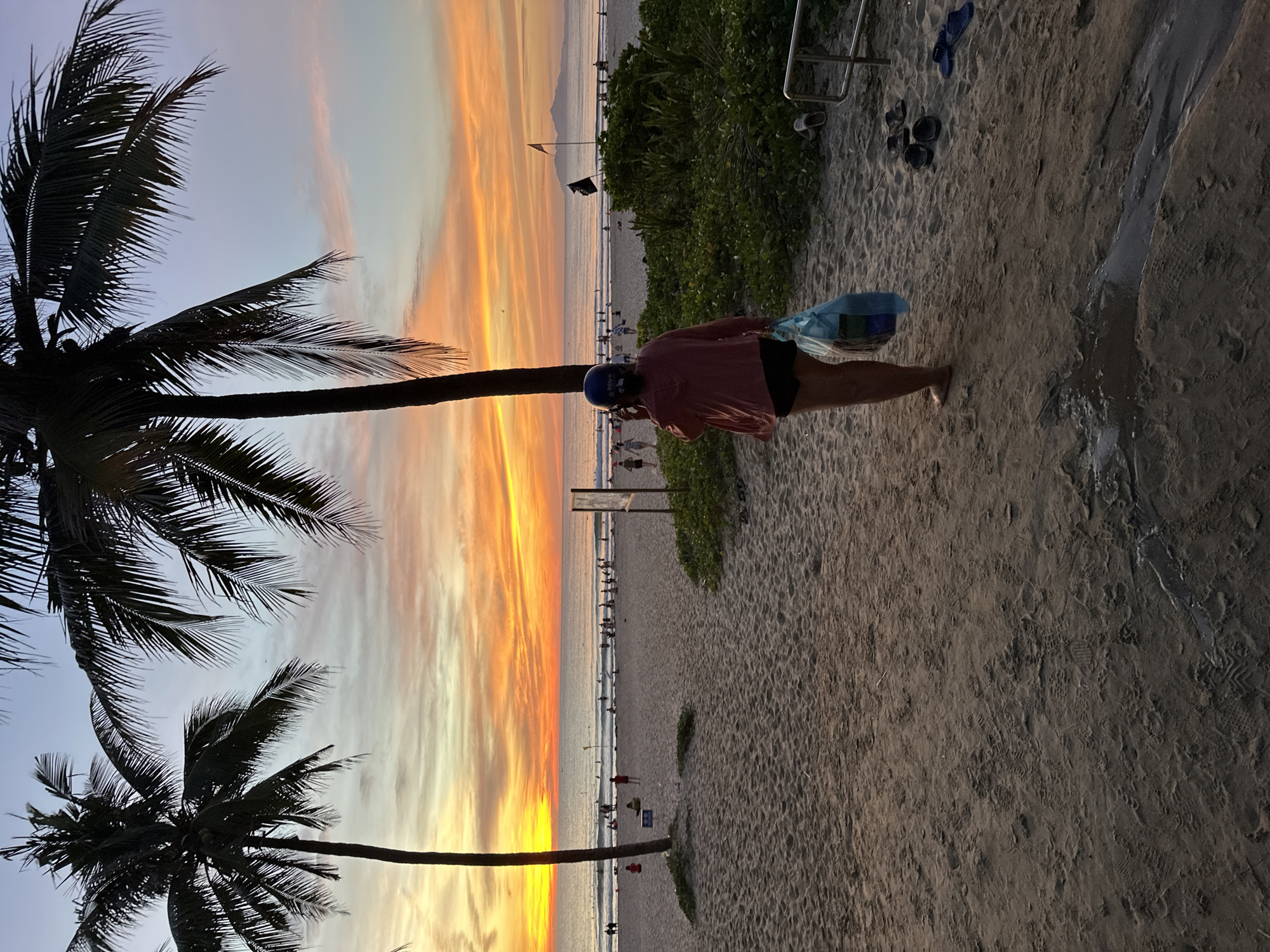 A person is standing on a sandy beach at sunset, holding a blue plastic bag. The person is wearing a pink top and dark shorts, with a blue helmet on their head. In the background, the ocean is visible with waves and a few surfers. The sky is filled with vibrant orange and yellow clouds. Two palm trees are in the foreground, silhouetted against the colorful sky. The beach appears to be populated with other people in the distance, some walking and some in the water. This scene is characteristic of My Khe Beach in Da Nang, Vietnam, a popular destination known for its beautiful sunsets and recreational activities.