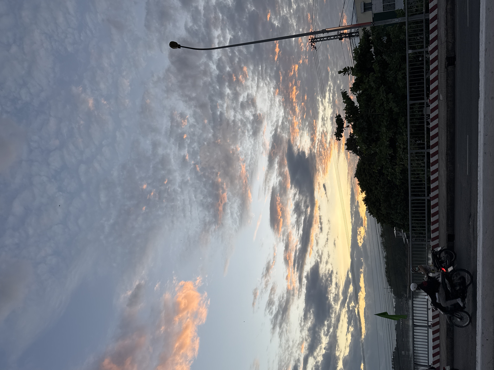 The image depicts a sunset scene over a bridge in Nha Trang, Vietnam. The sky is filled with a mix of light blue and gray clouds, with pockets of orange and pink light filtering through, indicating the sun is low on the horizon. A tall, dark street lamp stands on the right side of the frame, silhouetted against the sky, with power lines visible near its base. Below the sky, a dense line of dark green trees runs along the embankment behind a metal railing that lines the bridge. On the bridge, two motorcycles are visible. The motorcycle in the foreground carries two individuals, both wearing masks. The rider is wearing a white helmet and a dark top, while the passenger wears a patterned dress. Another motorcycle is partially visible behind them. The bridge itself has a white and red striped border along its edge. A green flag is attached to a pole on the left side of the bridge, near the railing. In the background, a body of water can be seen, with some structures and distant foliage on the opposite bank. The overall scene captures a moment of urban life during sunset, with elements of transportation, nature, and infrastructure present.