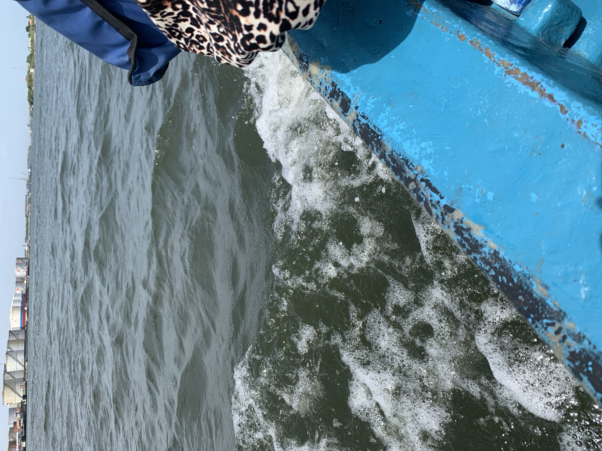 The image shows a view from a blue boat moving through water. White foam and bubbles are visible where the boat cuts through the water. Parts of a building can be seen in the distance, indicating a location near a shoreline or port in Ibasa, Nigeria. A piece of clothing, blue and leopard-print fabric, sits on the edge of the boat.