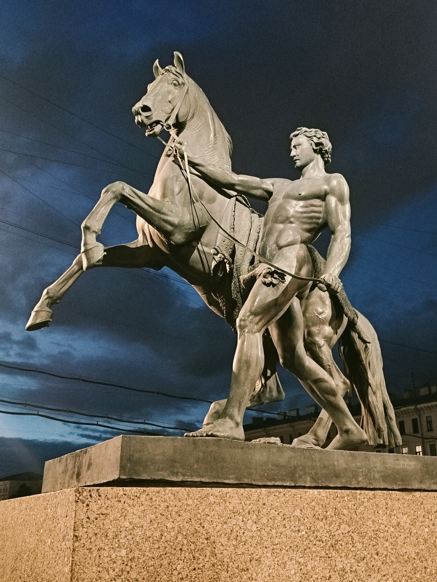 A low-angle shot reveals a bronze sculpture of a man and a horse, set against a twilight sky. The horse is rearing up on its hind legs, with the man standing beside it, gripping a rope or rein. Both figures are muscular and rendered in detail. The sculpture stands on a multi-tiered base, featuring a rough-textured lower layer and a smooth, rectangular upper layer. Overhead, wires cross the sky. In the background, there is a building with multiple windows. This is The Horse Tamers (also known as The Horsemen) statue in Saint Petersburg, Russia.