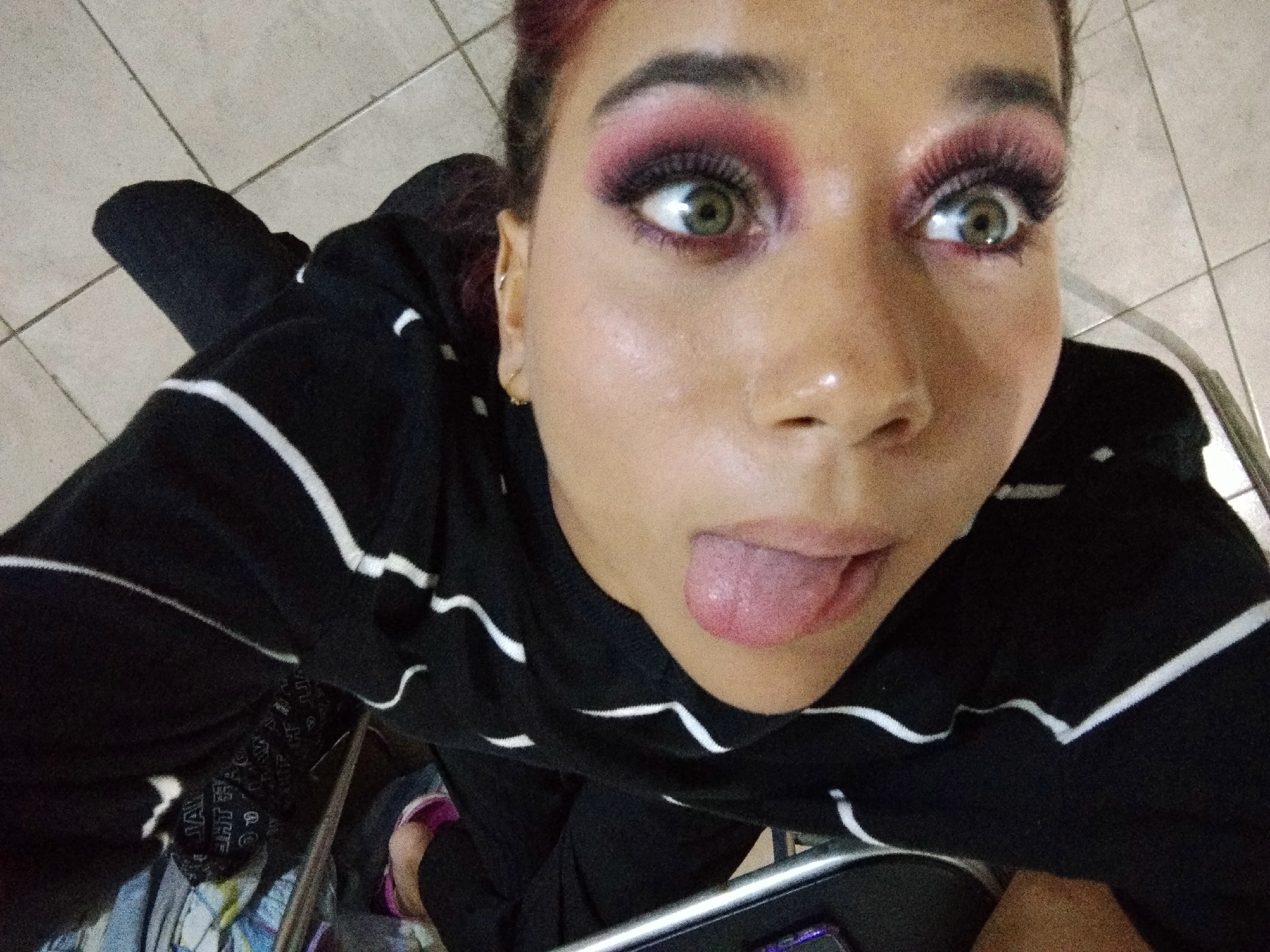 A young woman with pink hair and dramatic eye makeup sticks her tongue out while looking up at the camera. She is wearing a black and white striped sweater. Her eyes are green and she has large, false eyelashes. The background is a tiled floor.