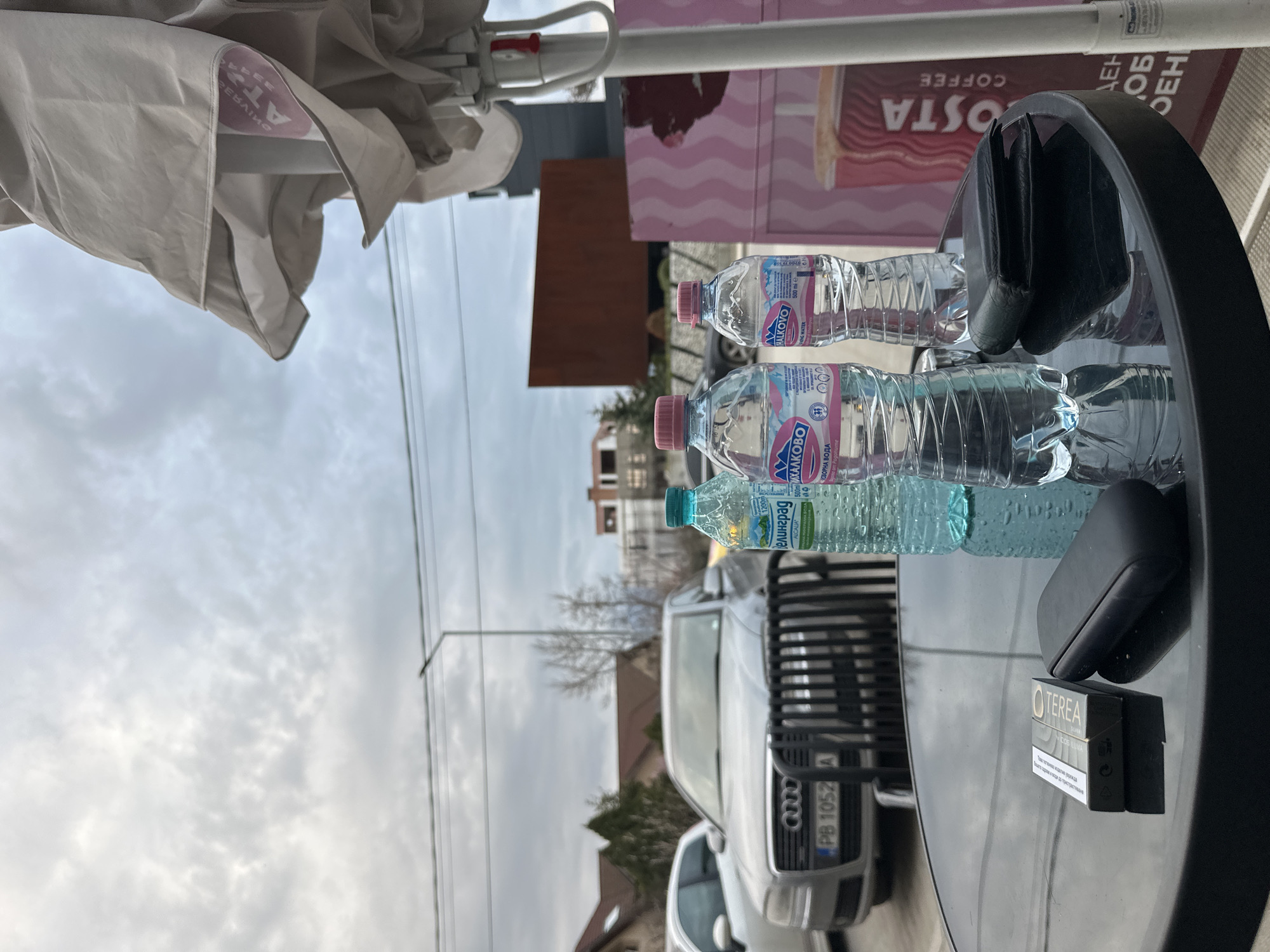 The image, rotated for easier viewing, depicts a table with various items on it, in what seems to be an outdoor seating area. There are three plastic bottles of water arranged in a row. The first two bottles are labeled "Михалково", while the third bottle is labeled "Велинград". Adjacent to the water bottles are two black objects and a pack of "TEREA" cigarettes. The table is black and glossy, reflecting the objects placed on it.
In the background, a white car is partially visible. The license plate reads "PB 1052 A". Behind the car are buildings and trees, suggesting an urban setting. The cloudy sky indicates overcast weather.
Parts of an umbrella are visible at the top left of the image, indicating this is an outdoor space. In the background is a Costa Coffee advert. The overall setting appears to be a cafe or restaurant with outdoor seating, likely located in the city of Plovdiv, Bulgaria, based on the water brands and license plate details.