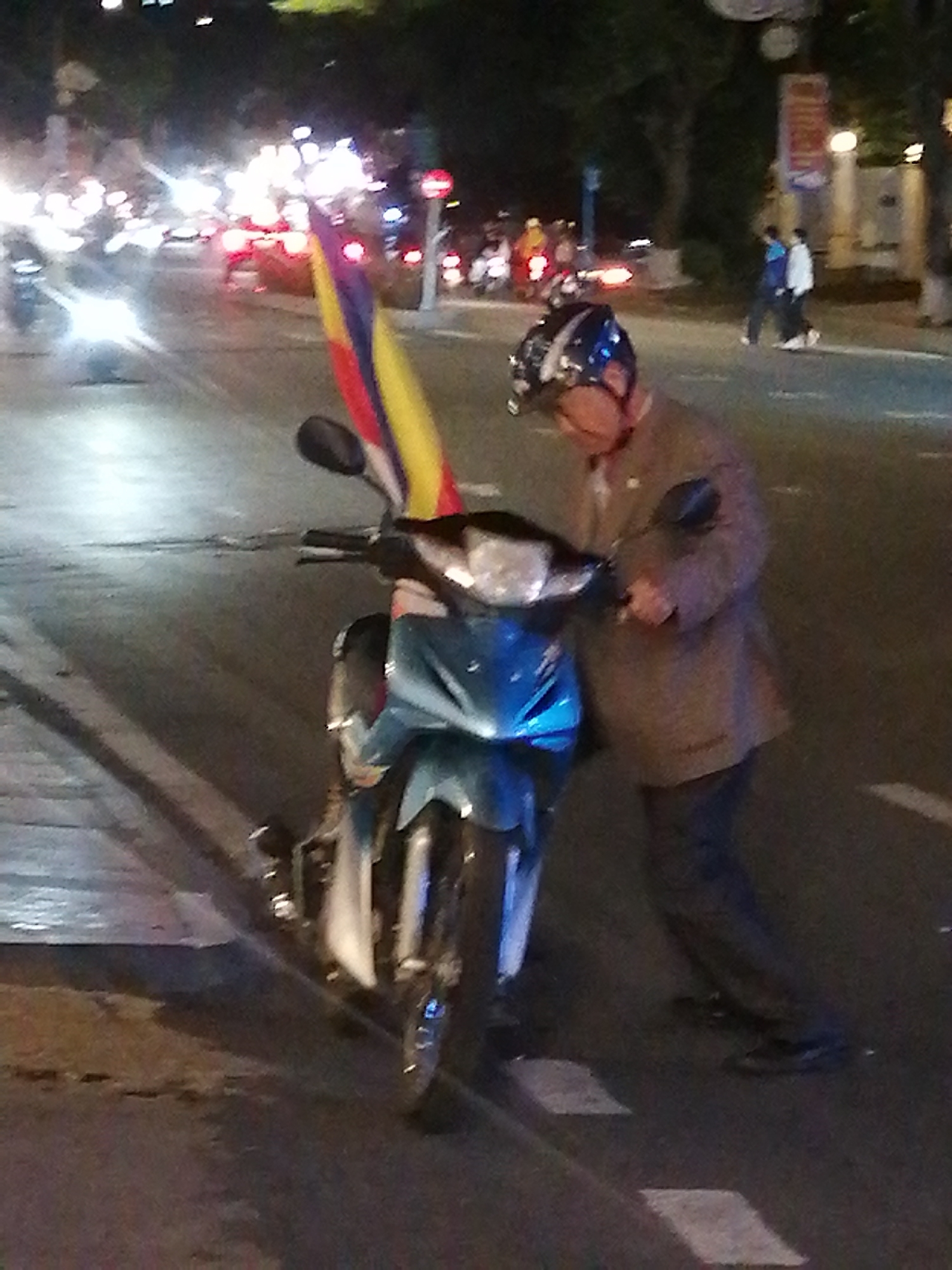 At night in Da Lat, Vietnam, a man wearing a helmet and jacket stands on a crosswalk next to a parked blue scooter displaying a vertically oriented flag. The scooter's headlight is on. Car headlights and taillights are visible in the background street scene. Two pedestrians are visible on the sidewalk in the background. Trees and buildings line the road, and a sign is visible.
