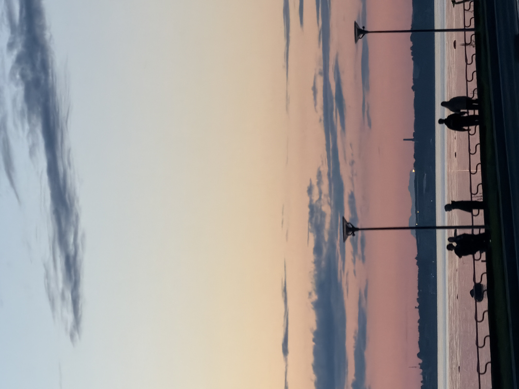 A group of people are silhouetted against a sunset sky on a waterfront promenade in Tallinn, Estonia. The sky transitions from a pale blue at the top to a soft orange and pink hue near the horizon, with scattered clouds. A body of water, likely the Baltic Sea, stretches across the mid-ground, reflecting the colors of the sky. A dark railing runs along the edge of the promenade, separating it from the water. Several lampposts are visible along the promenade, and in the distance, a silhouetted treeline and some structures are discernable on the opposite shore. The people, all appearing as dark figures, are standing and walking along the promenade, some in pairs and others individually. The scene appears to be a peaceful evening setting.