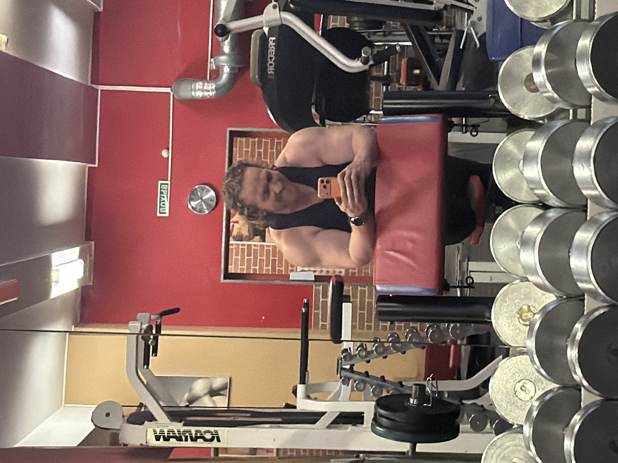The image depicts a gym interior in Saint Petersburg, Russia, captured as a mirror reflection. In the center, a muscular man with light-brown curly hair and a short beard, wearing a black tank top and a watch on his left wrist, smiles faintly while looking down at an orange smartphone. He holds the phone with both hands, resting his forearms on the red padded armrest of a bicep curl machine.
In the reflected foreground, a rack of silver dumbbells is visible, some marked with the number "8" on their ends. Behind the man, other gym equipment is reflected: a white multi-station machine with the mirrored text "WANNAI" on its side, and a black Precor machine (identified by its mirrored brand name "ЯОССЭЯЧ"). A rack of smaller chrome dumbbells is also visible behind the multi-station machine.
The actual gym environment, seen in reflection, features red and beige-yellow painted walls and a white ceiling with integrated fluorescent light fixtures. A green sign with white Cyrillic text "ВХОД" (Entrance) is mounted on a red wall, positioned above a small white circular object and to the right of a silver circular wall clock. A large silver ventilation duct runs along the upper right portion of the room. A framed picture, possibly of a human torso, is visible on the beige-yellow wall, and a faux brick pattern frames an opening on the right side.