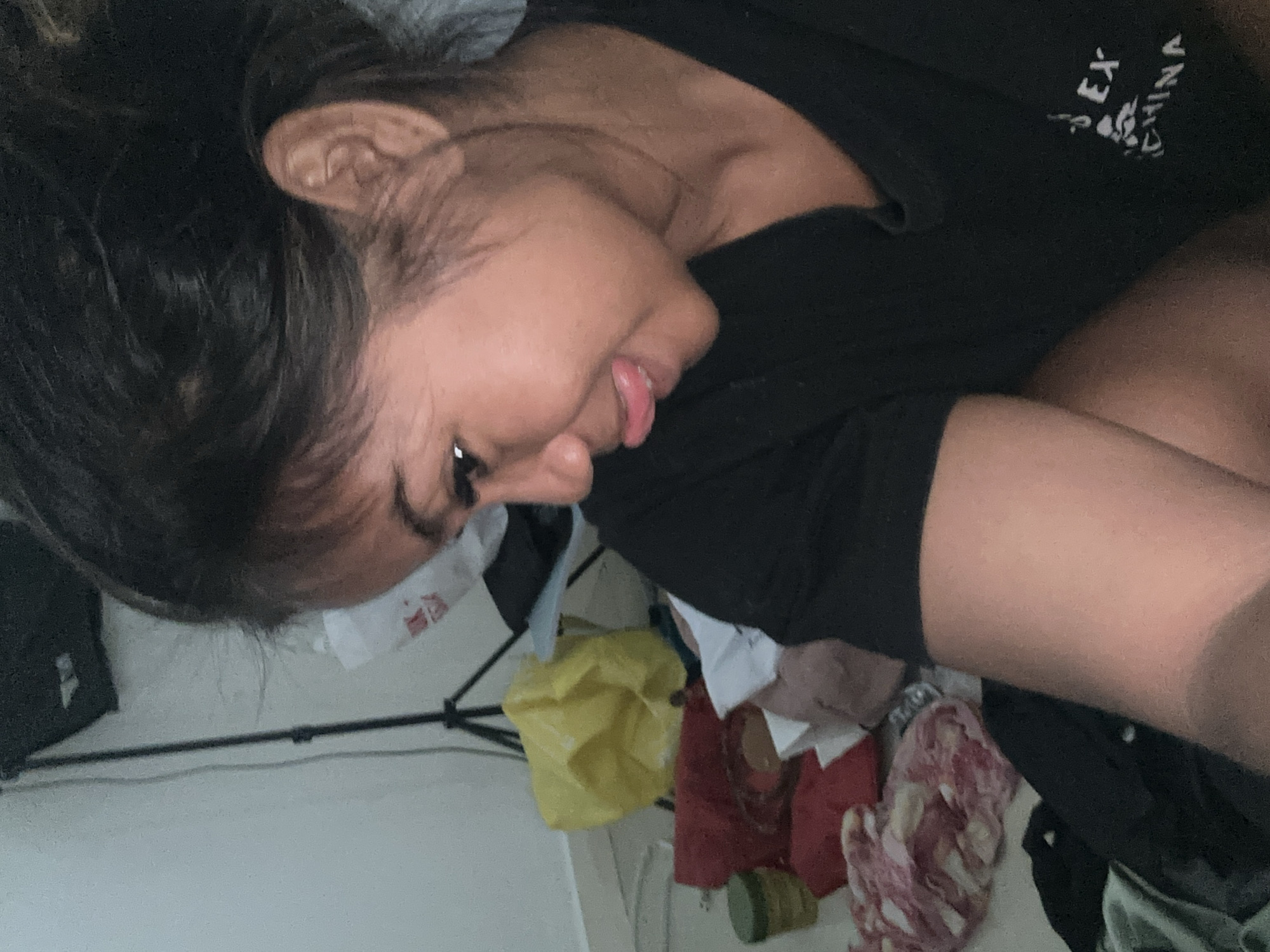 A young woman with dark hair pulled back is shown in a close-up profile shot, sticking her tongue out to the side. She is wearing a black t-shirt with white text that reads "SEX CHINA." The background is cluttered with various items, including a yellow plastic bag, red fabric, a green jar, and other unidentifiable objects. A black tripod is visible in the upper left corner. The lighting suggests an indoor environment, possibly a room with minimal light. The woman's skin tone is a medium brown. The overall scene appears to be candid and informal, with the woman in a playful pose.
