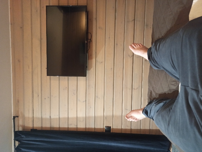 The scene is an indoor environment in Nizhneye Zadolgoye, Russia, captured from a first-person perspective of an individual lying supine. The lower torso, legs, and bare feet of a person wearing dark grey casual trousers are visible in the foreground. The person rests on a brown, quilted surface. Ahead is a wall clad with light-toned horizontal wooden planks. A dark blue pleated curtain hangs from a black rod on the left side of the wall. A black flat-screen television is mounted on the wooden wall to the right, displaying a dark, unlit screen with visible cables underneath.