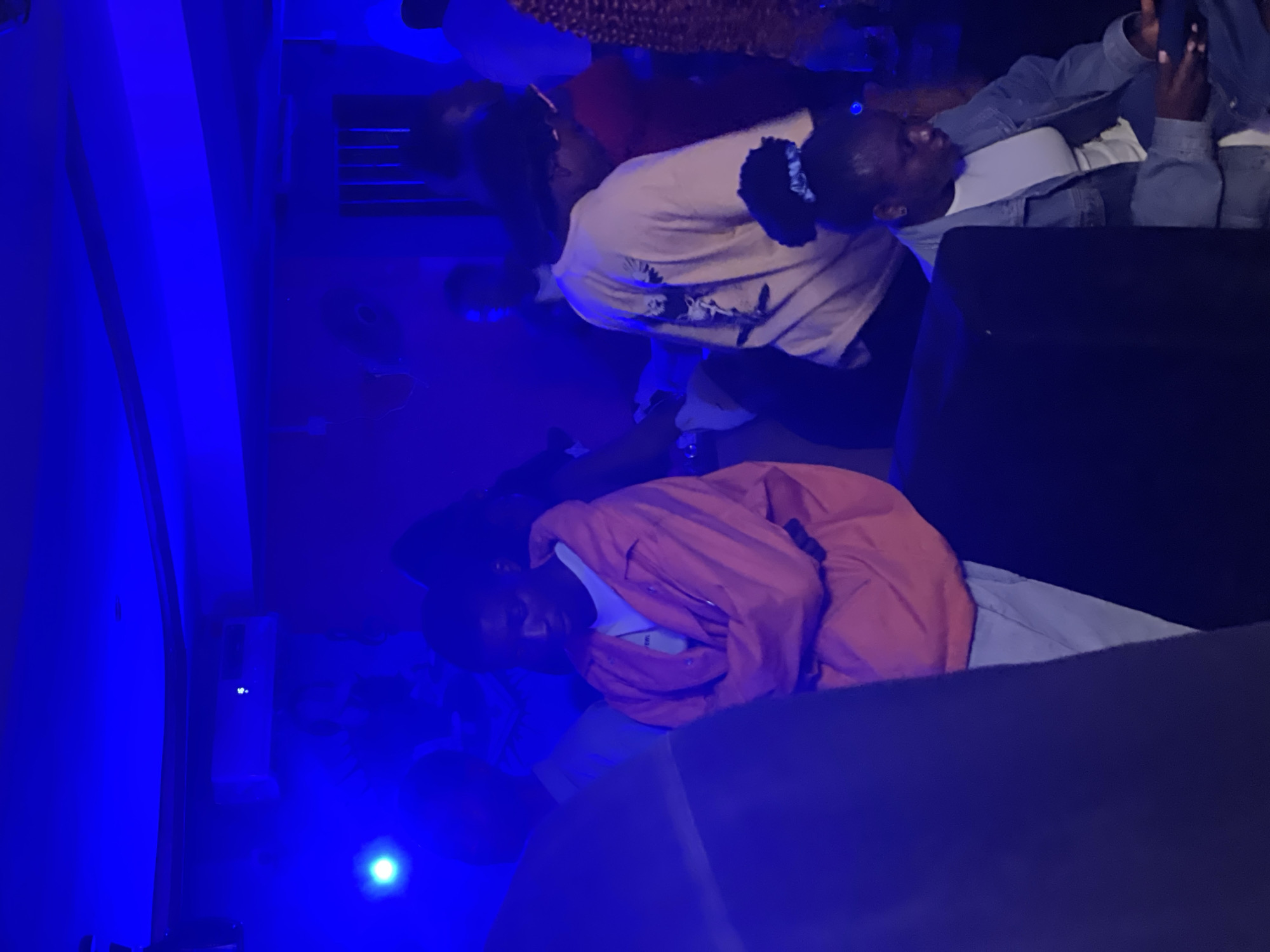 This is a medium shot, indoor photograph taken at night, in the city of Durumi, Nigeria. The scene appears to be a dimly lit gathering, possibly a party or social event, with heavy blue lighting dominating the environment.
In the foreground and slightly to the left, a person is visible from the chest down, wearing a dark-colored garment that obscures much of their form. Their presence suggests they are standing or walking through the space.
To the right of center, a young man is the primary subject. He is wearing a white t-shirt under an open, bright orange jacket. His arms are crossed across his chest, and he is looking down with a pensive expression. He appears to be standing and engaged in the event.
Further to the right, partially obscured, is another young woman. She is wearing a denim jacket over a white top and has her hair tied up with a blue scrunchie. She is seated and looking down at a dark object in her hands, possibly a smartphone.
In the background, other individuals are present but largely indistinct due to the low light and the blue ambient illumination. There are hints of furniture or structures within the space, and on the back wall, there is a visible air conditioning unit. The overall atmosphere is that of a casual, late-night social gathering in an urban setting in Durumi, Nigeria.