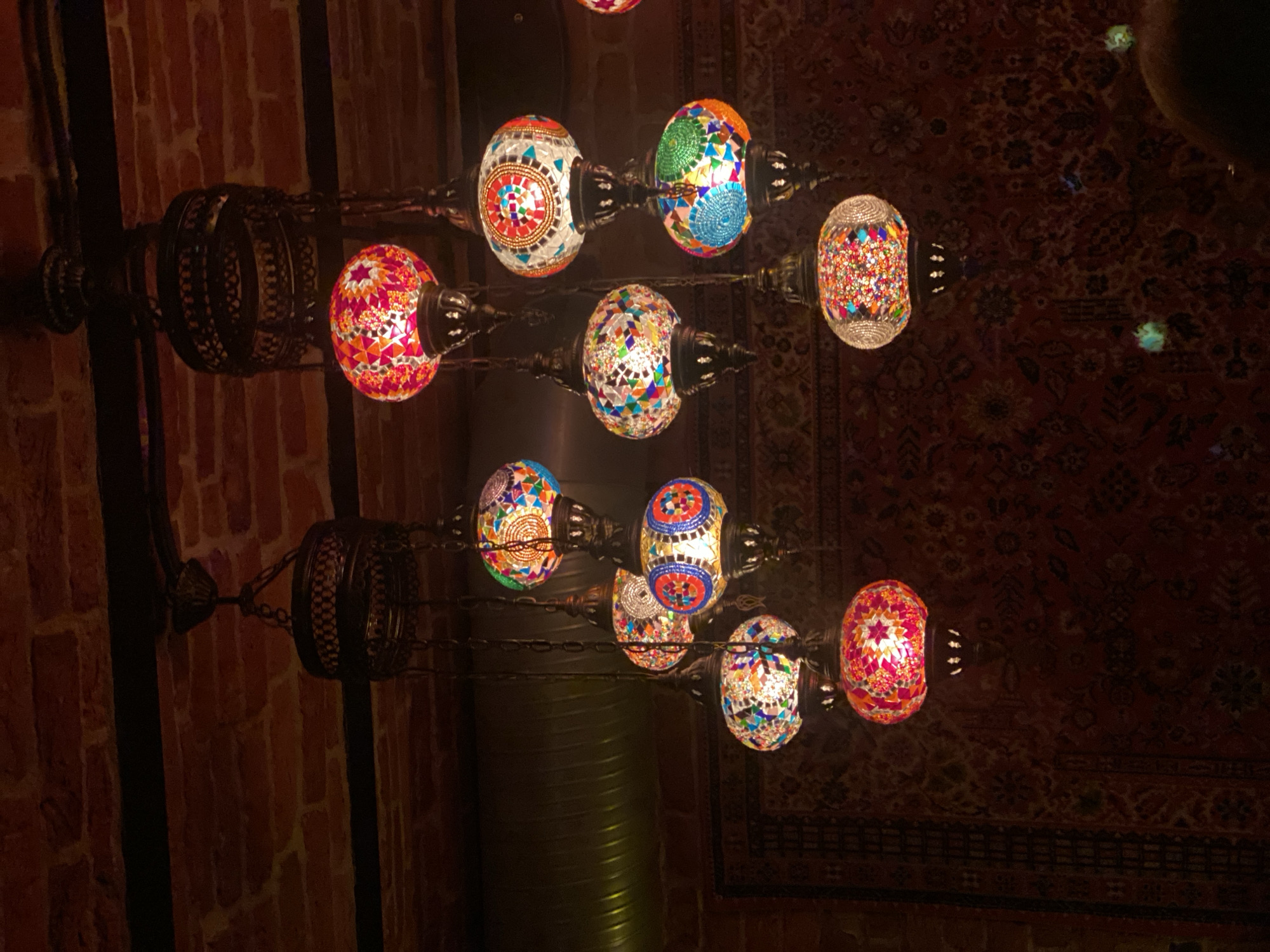 The image depicts multiple mosaic glass lamps suspended from a ceiling constructed of exposed red brickwork and dark horizontal beams. A dark, corrugated metal ventilation duct is visible on the upper left ceiling. Each lamp, varying in size and shape (predominantly spherical or gourd-like), is composed of numerous small, irregularly cut glass pieces in colors including red, orange, yellow, green, blue, and white, illuminated internally. The lamps are attached to dark metal fixtures and chains, hanging at different lengths and forming clusters. The background features a dark red, intricately patterned rug or tapestry. A partial view of a human head, with dark hair, is present in the bottom right corner. No specific actions or interactions are discernible. The depicted location is Saint Petersburg, Russia.