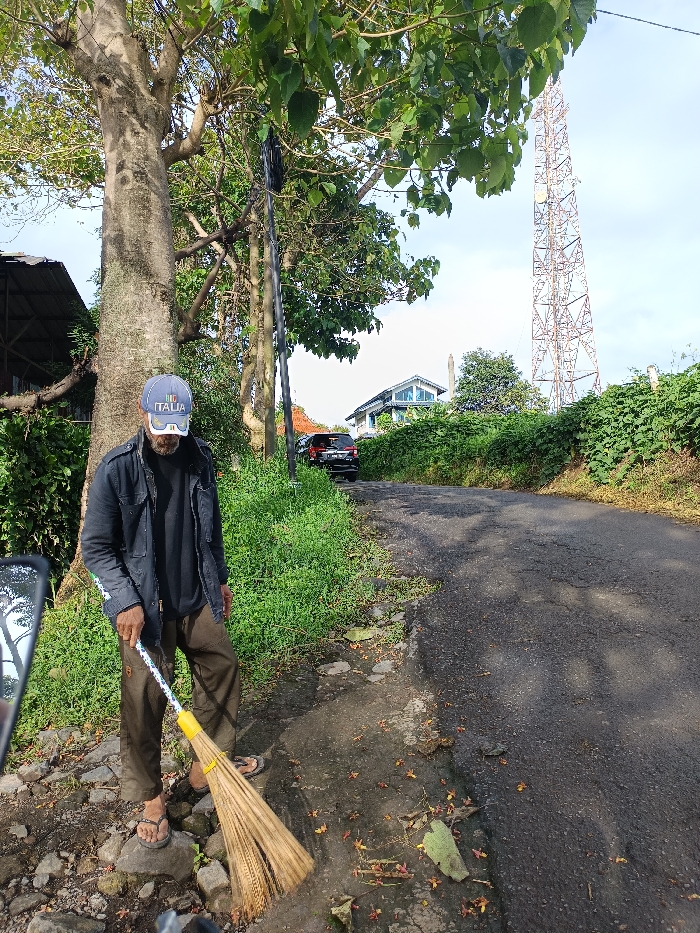 A man stands near a tree, wearing a dark jacket, brown pants, sandals, and a cap with "Italia" on it. He holds a broom. He is on the side of a paved road with patches of exposed earth and scattered leaves. In the background, a car is parked on the road near a house, and a tall metal tower rises against the sky. Green vegetation lines the sides of the road.