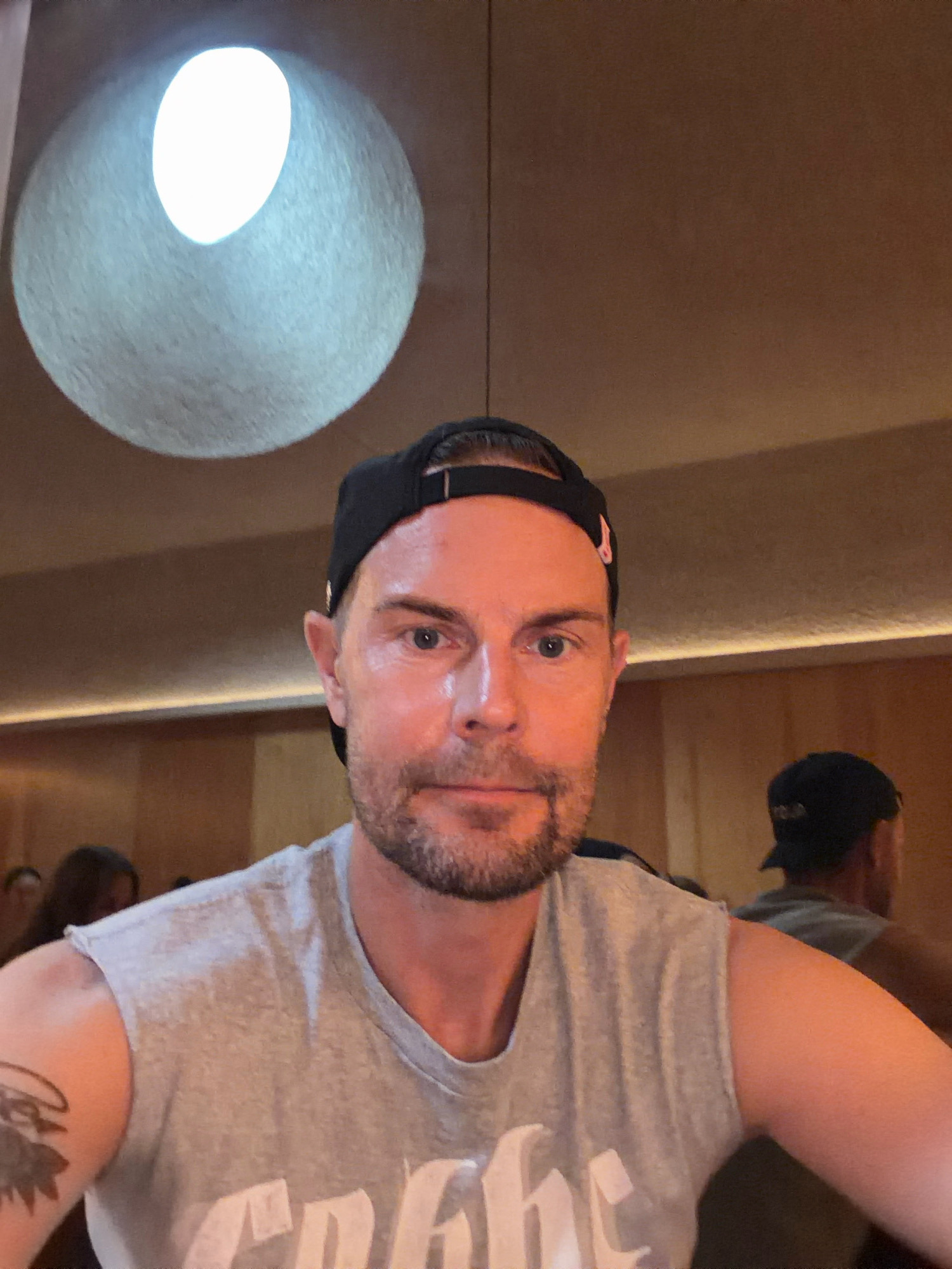 A man wearing a gray sleeveless t-shirt and a black baseball cap is pictured from the chest up, indoors. He has a short beard and light-colored eyes, and his left arm displays a tattoo. In the background, another person wearing a black baseball cap can be seen, as well as other blurred individuals and a large, circular light fixture on the ceiling. The walls are a warm brown color, with a strip of light running horizontally along the lower portion. The scene appears to be in a restaurant or bar.