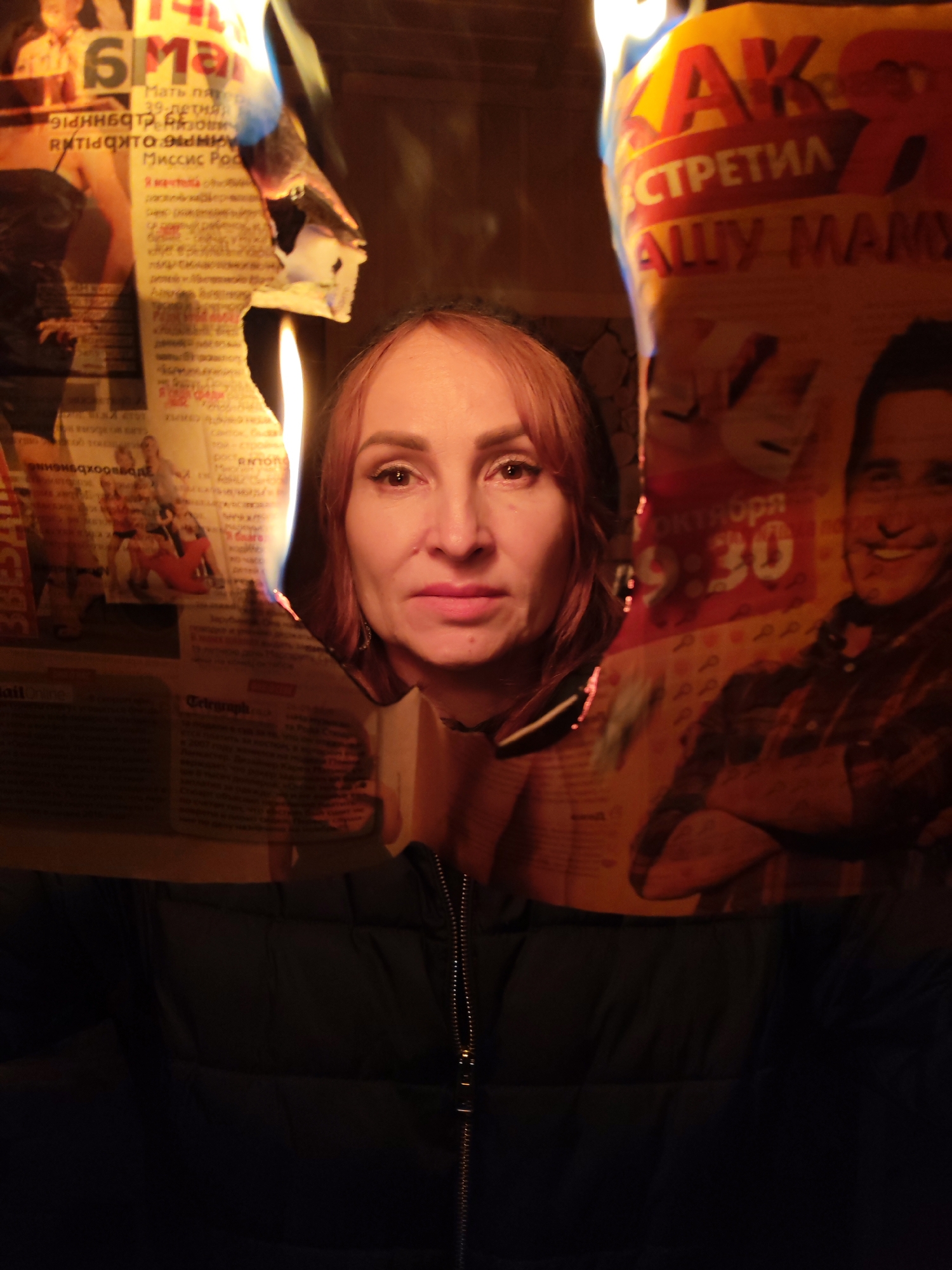 A woman stands facing the camera, her head framed by a burning newspaper. The newspaper has various text and images, including the words "Мать пятер", "Миссис Рос", "СТРЕТИЛ АШУ МАМУ", and "Октября 9:30". The woman wears a dark jacket with a visible zipper. Flames are visible on both sides of her head. The location is Unknown, Unknown.