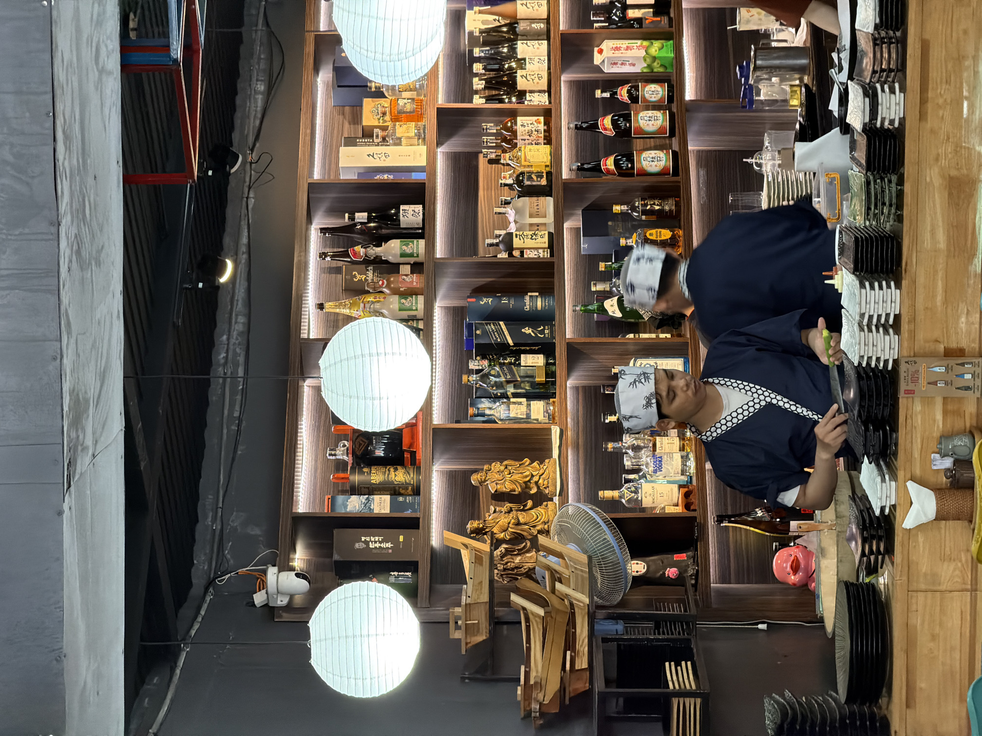 In Nha Trang Ward, Vietnam, a bar or restaurant scene is captured. Two individuals, likely chefs or bartenders, are present. One, wearing a dark blue traditional Japanese robe with white trim and a white headband, stands behind a wooden counter, appearing to prepare food. He is holding a green vegetable and a knife. To his right, another individual in a similar robe is visible from the back, also behind the counter.
The environment features a wall lined with shelves displaying a variety of bottles, predominantly spirits, and some boxed items. Notable brands include Macallan, Absolute Vodka, and Glenfiddich. There are also decorative wooden boat models and figurines on the shelves. Hanging from the ceiling are two large, white, round lanterns. The counter area is organized with stacks of plates, bowls, and serving dishes, along with some kitchen equipment like a fan and a metal pitcher. A pink novelty item with eyes is visible on the counter. The overall lighting is dim, with warm light emanating from the shelves.