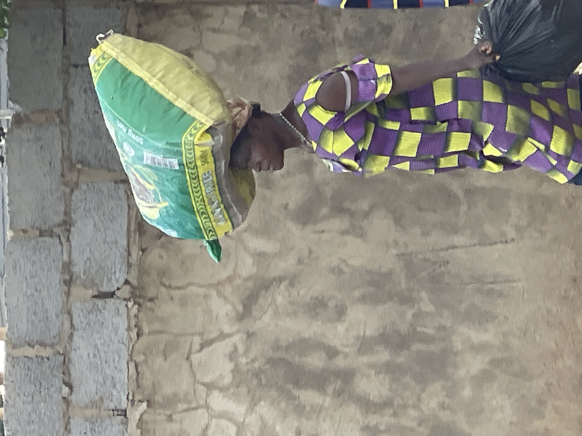 An African woman is carrying a large green and yellow sack on her head in Abuja, Nigeria. She has a black trash bag in her left hand and is wearing a purple and yellow checkered shirt. The background includes a concrete block wall and a textured, plastered wall. The setting appears to be a construction site.