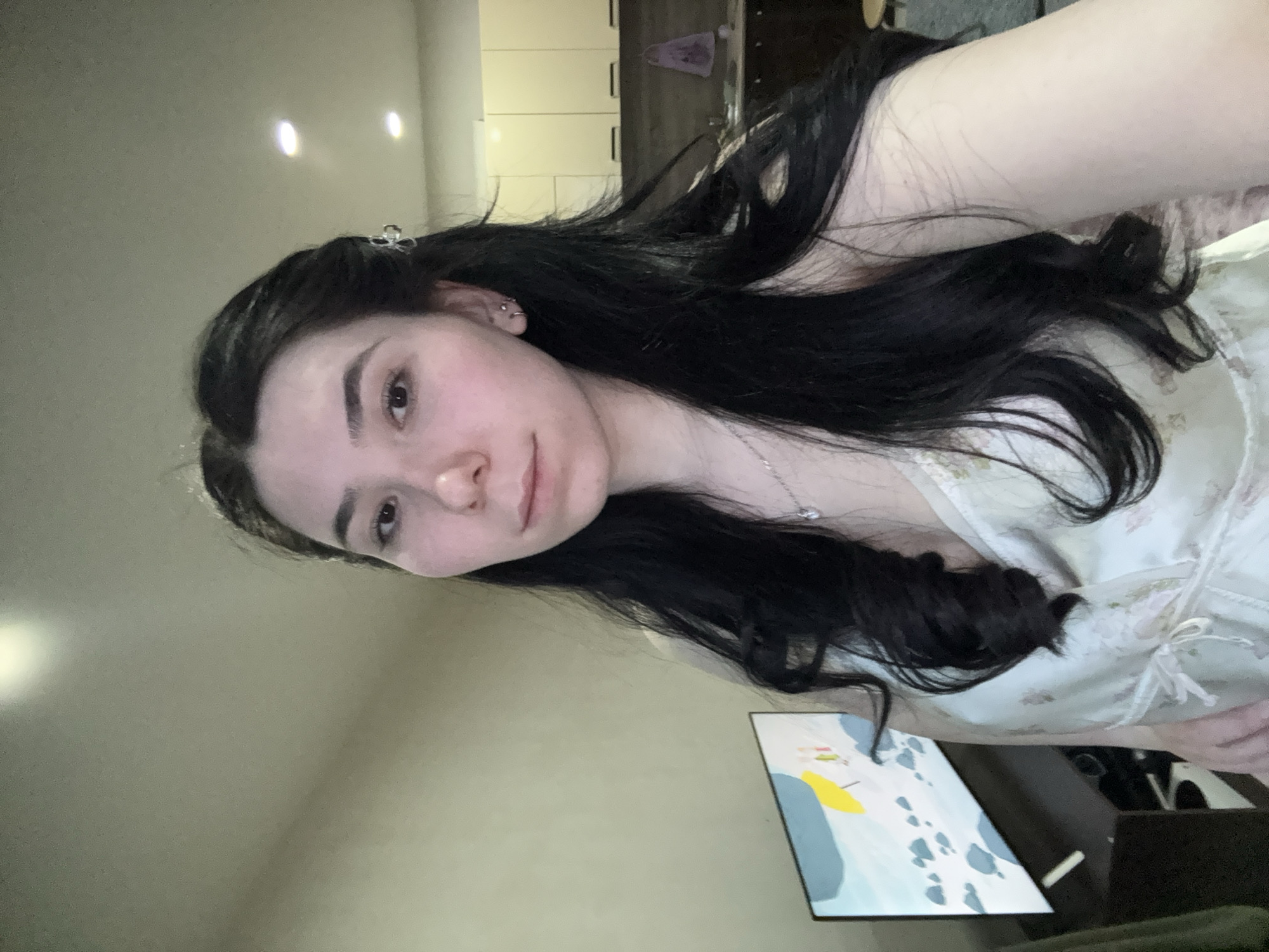A young woman with long, dark hair is pictured indoors. She is wearing a light-colored, patterned top and a delicate silver necklace. Her hair is styled with a small clip on one side. She appears to have multiple piercings in her ear. In the background, there is a glimpse of kitchen cabinets and a piece of abstract art. There's no indication of the city Izhevsk, Russia.