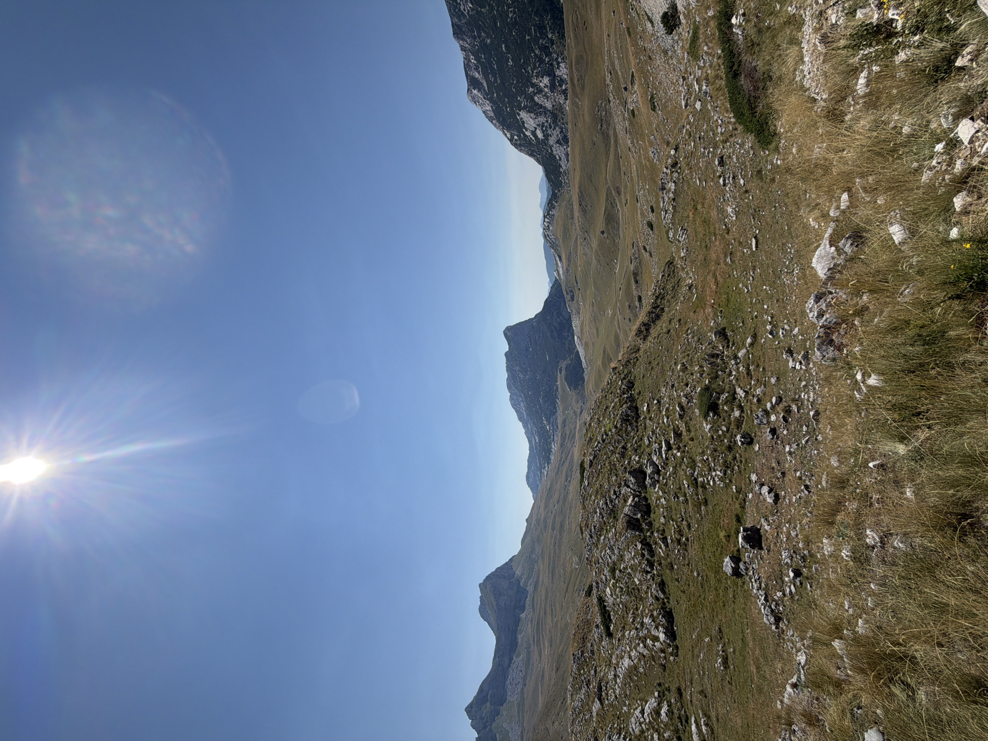 The image depicts a mountainous landscape under a clear blue sky. The sun is prominently visible in the upper left corner, emitting bright rays and causing a lens flare. The terrain is characterized by rolling hills covered in dry grass and scattered rocks. In the distance, several mountain ranges are visible, with varying textures and shades of grey and green. The overall environment appears arid and somewhat desolate, with no visible signs of human habitation or activity. The location is identified as Dubrovsko, Montenegro.