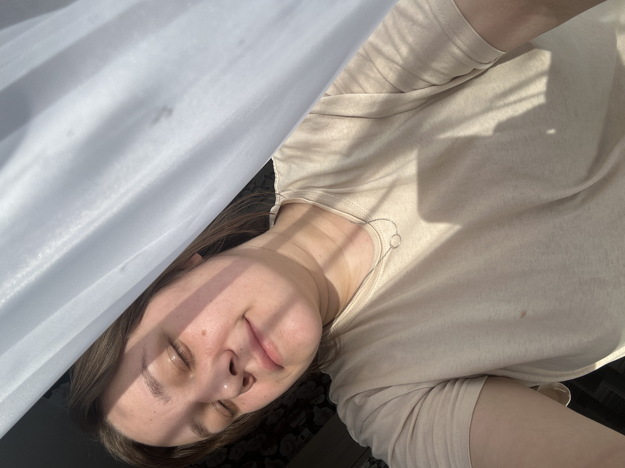 A woman is shown indoors, likely near a window due to the natural light and the presence of a white curtain. She appears to be lying down, possibly on a bed or couch. She has her eyes closed and is wearing a light-colored t-shirt. The location is unknown.