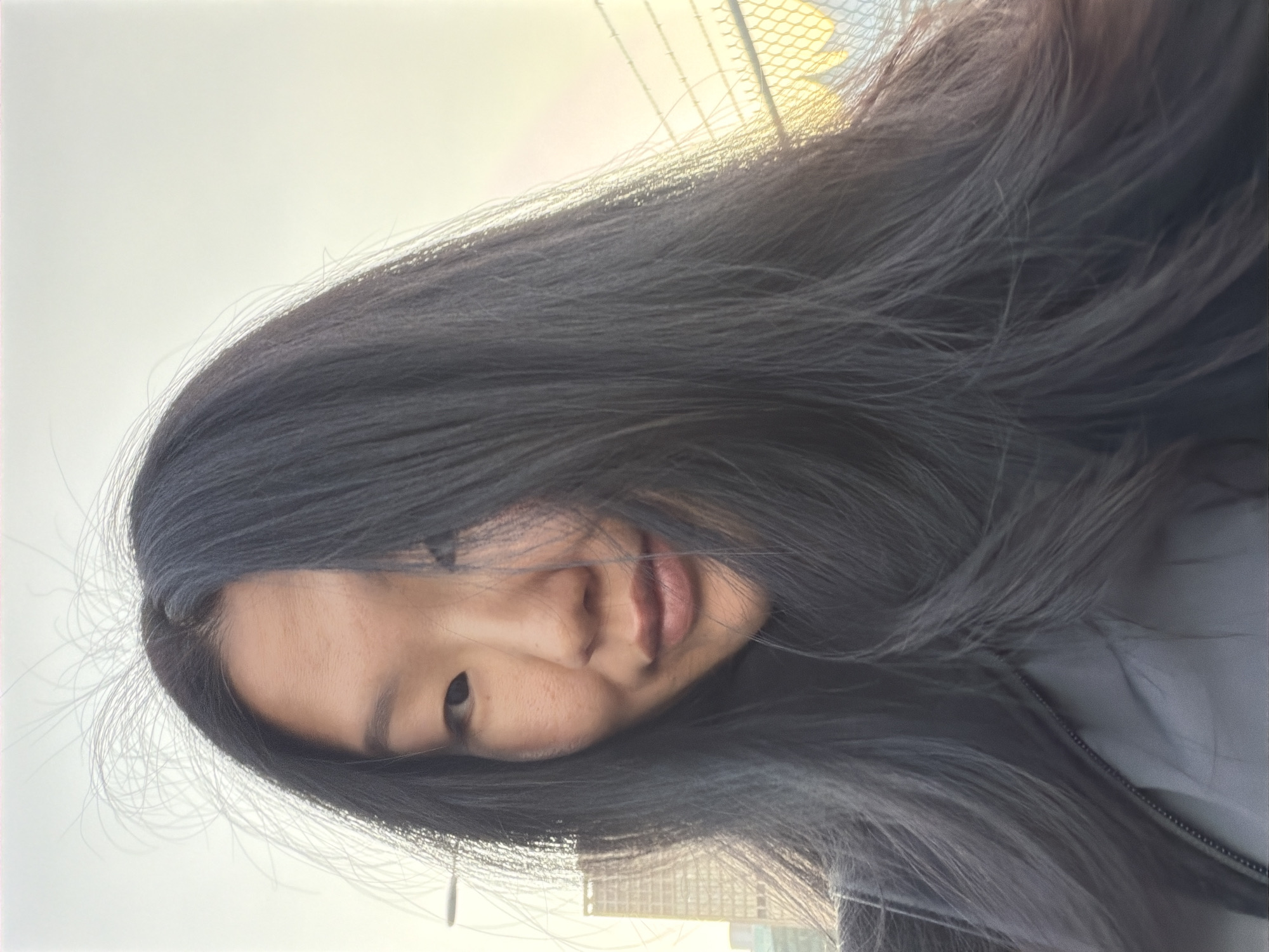 The image is a close-up selfie of a woman with long, dark hair, taken outdoors. She is wearing a grey zippered jacket. The background shows a hazy sky, a tall building, and a chain-link fence. The lighting suggests it is either dawn or dusk. The photograph is not located in Vancouver, Canada.