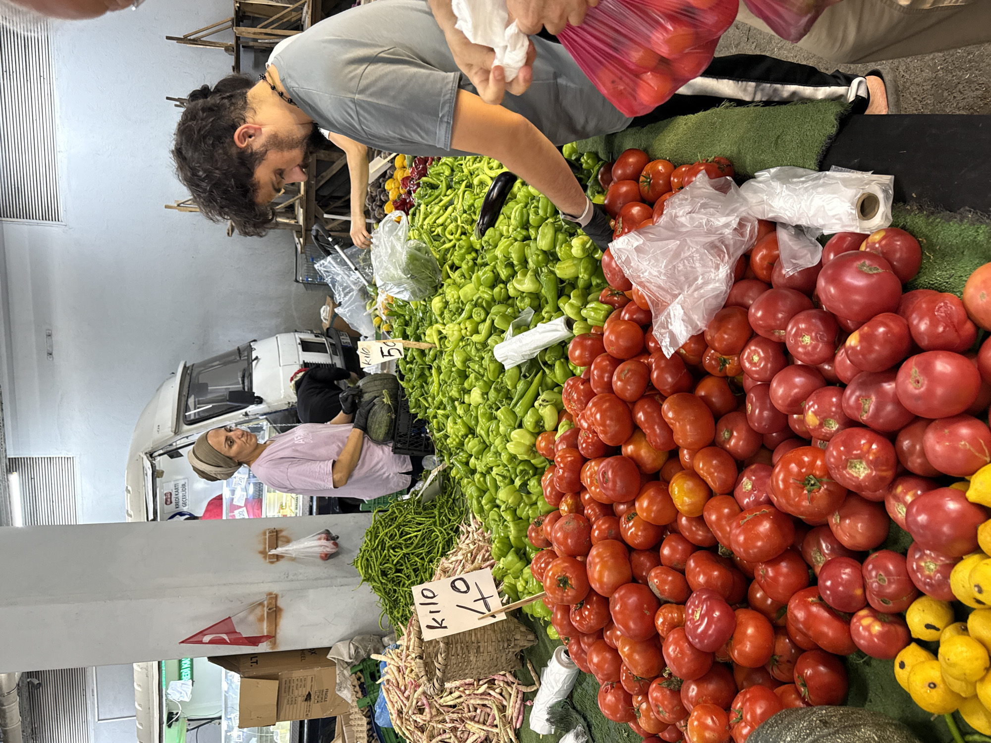 In Kadıköy, Turkey, a bustling open-air market is depicted. A woman wearing a headscarf and a pink t-shirt stands behind a large display of fresh produce, primarily red tomatoes and green peppers. She is wearing black gloves and appears to be attending to the vegetables. Beside her, a man with curly hair, also wearing black gloves and a grey t-shirt, is seen reaching into a pile of green peppers, possibly selecting them. In the background, a white food truck is visible, with signage indicating it is a vendor. Various other vegetables, including long green beans and yellow fruits, are also displayed on tables and in baskets. The market appears to be busy, with the presence of both vendors and customers, suggesting a lively commercial environment.