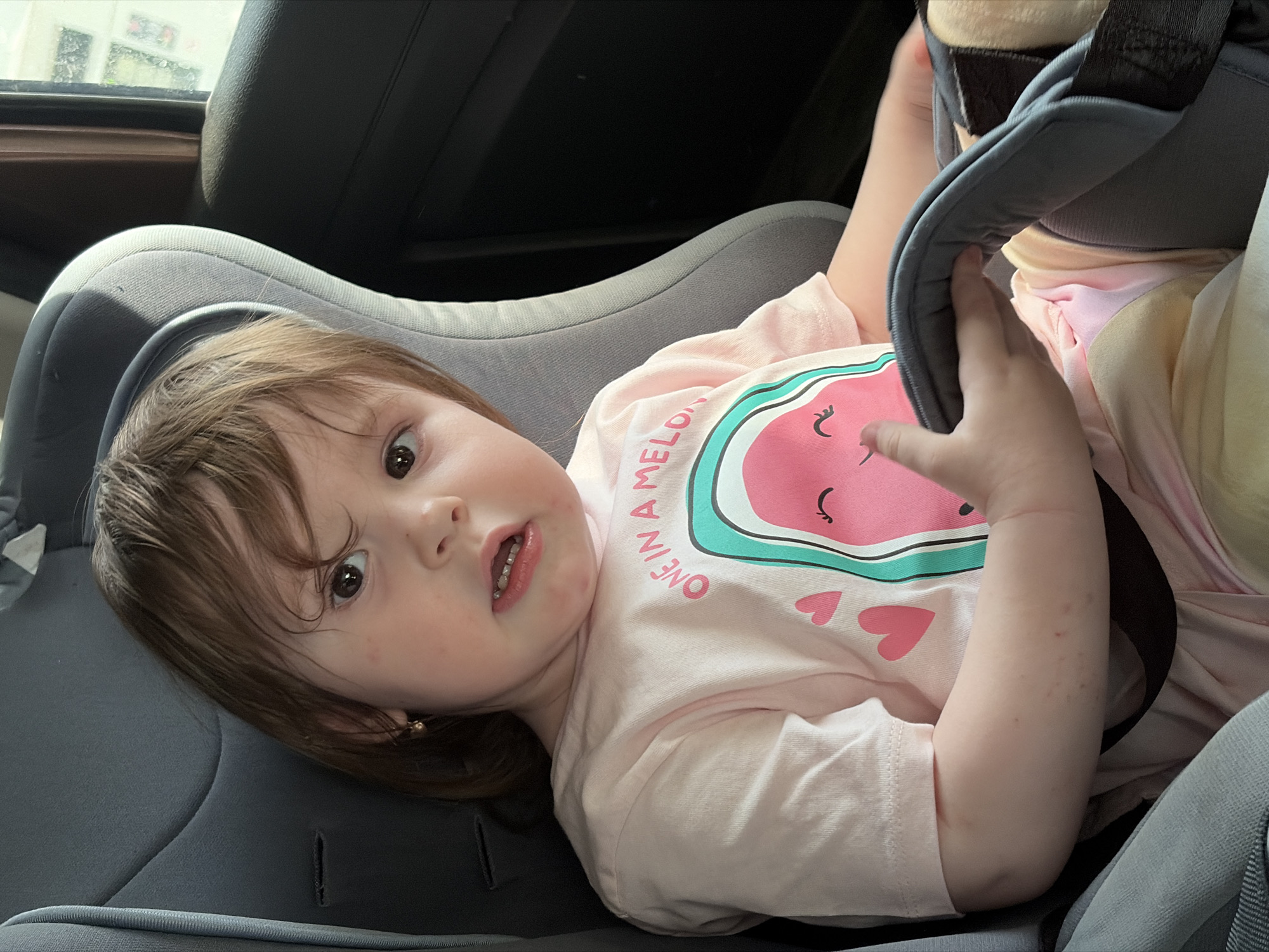 A young child with light brown hair is secured in a gray car seat. She wears a pink t-shirt with a watermelon design and the words "ONE IN A MELON" printed on it. The car's interior includes a black headrest and partially visible window. The location is unidentifiable as the image does not contain any contextual evidence pointing to the city of Chaikovsky, Russia, or any recognizable landmarks.