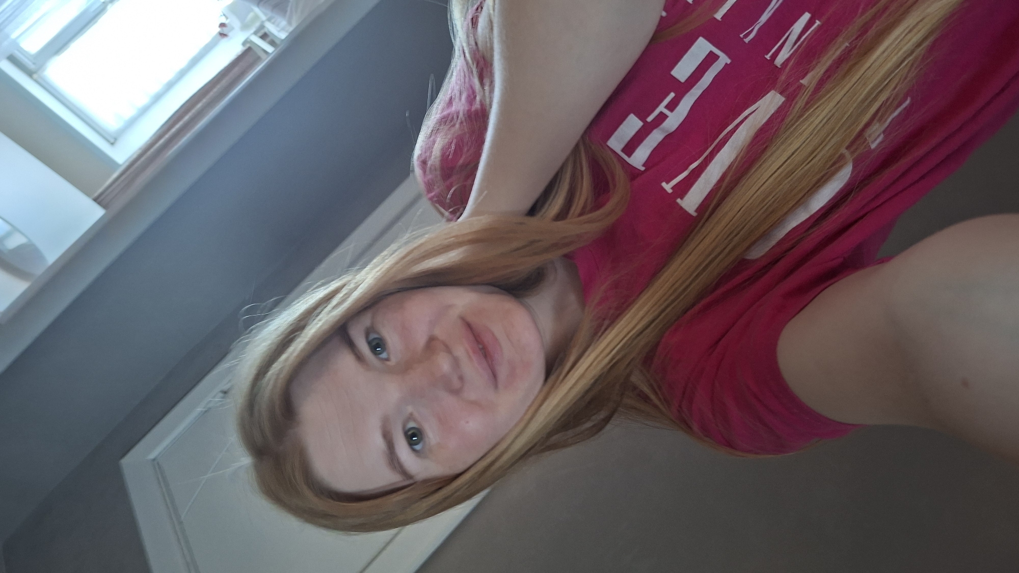 A young woman with long blonde hair and fair skin is taking a selfie. She is wearing a bright pink t-shirt with white lettering. Her head is tilted, and she is smiling slightly. Her right arm is raised, holding the camera. The background is a mix of a gray wall and a white door or frame. The image appears to be taken indoors, possibly in a bedroom or living area. There is a window visible in the upper right corner of the image, with sheer curtains. The lighting suggests it is daytime. The location is not specified in the image or accompanying text.