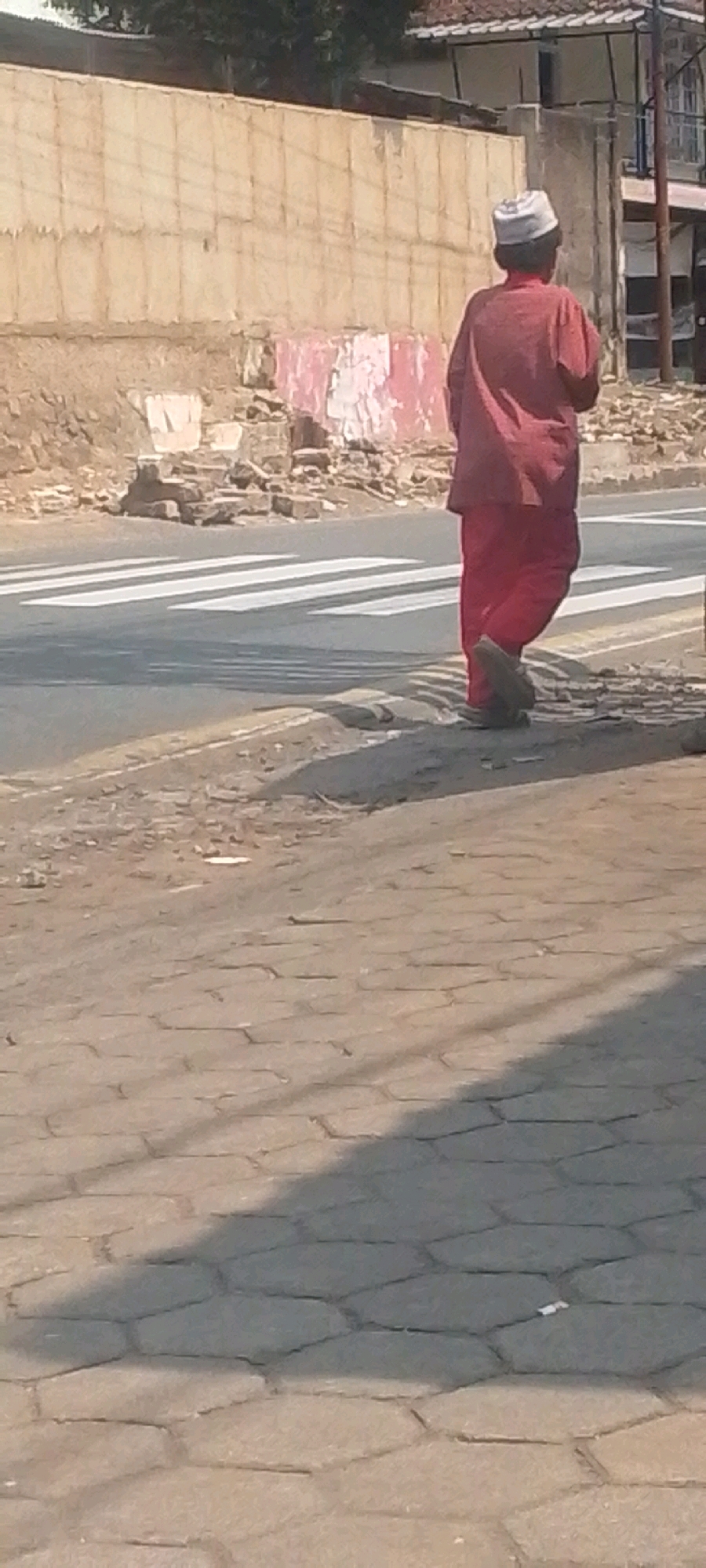 In Sirnagalih, Indonesia, a person is walking across a pedestrian crossing. The individual is wearing a red garment and a white cap. The environment is urban, with a road, sidewalk, and a wall visible. The wall appears to be made of concrete blocks and has some signs of damage. There is also a pile of debris to the left of the wall, suggesting construction or demolition. The ground is paved with cobblestones. The lighting suggests it is daytime, with shadows cast on the ground.
