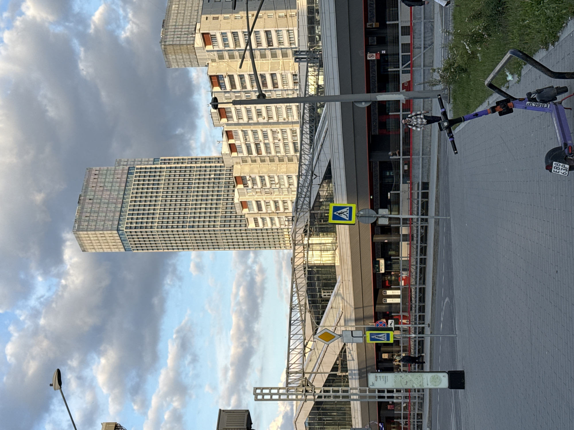 The image captures an urban street scene in Moscow, featuring a blend of modern and older architecture under a partly cloudy sky. In the background, a tall, modern skyscraper with a grid facade stands alongside a multi-story, beige-colored apartment building with numerous windows and balconies.
At street level, a wide paved area, possibly a sidewalk or plaza, leads to a road with white dashed lines. A complex metal structure, potentially an overpass or part of a station entrance, spans across the middle ground. Various street furniture is present, including streetlights and several traffic signs. Visible signs include a yellow diamond indicating a main road, blue pedestrian crossing signs (white person walking on a blue triangle within a blue square), and a red circular sign with a blue diagonal line, likely signifying "no stopping" or "no parking."
A purple electric scooter is parked on the right side of the frame, leaning against a metal bike rack. The scooter bears the alphanumeric codes "EBB 63P" and "EBB863P" on its rear and front. A person wearing a plaid shirt is visible near the scooter. Further down the street, another individual can be seen walking.
The sky is characterized by a mix of grey clouds and patches of blue, suggesting a partly cloudy daytime. The lighting appears soft, possibly indicating late afternoon. The scene depicts a functional and busy urban environment with contemporary public transport infrastructure and residential buildings, characteristic of a major city like Moscow. On a dark building facade on the right, small, indistinct signage includes what appears to be a "no smoking" symbol.