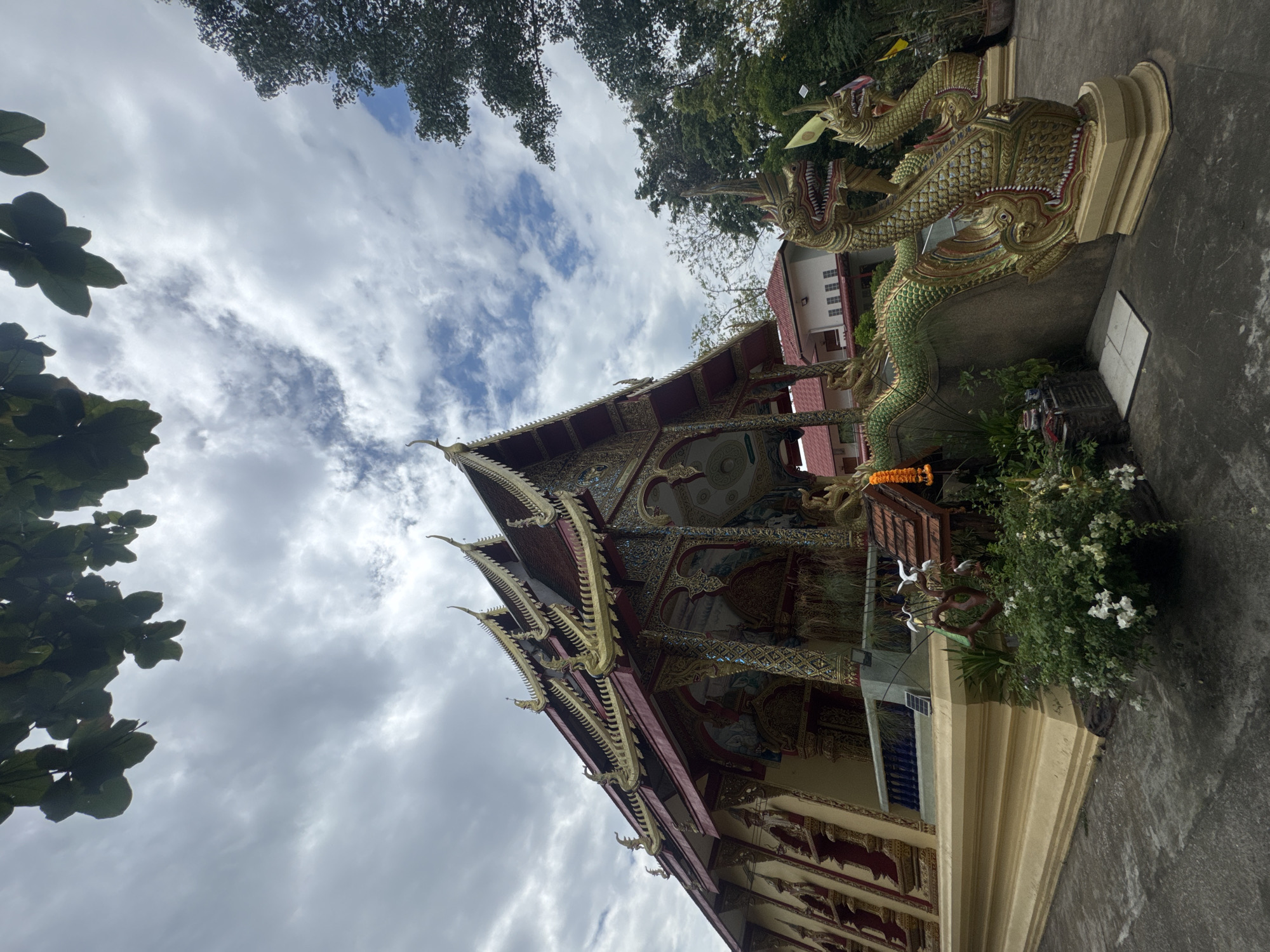 An eye-level shot shows a Buddhist temple in Ban Sai Kham, Thailand. The angle is unusual. It's oriented 90 degrees to the left.
The temple's ornate roofline is adorned with carvings. A large golden dragon statue is adjacent to the building. A garland hangs from the dragon's neck. A flowering bush grows at the base of the statue. The sky is overcast with puffy clouds. Trees frame the scene.
