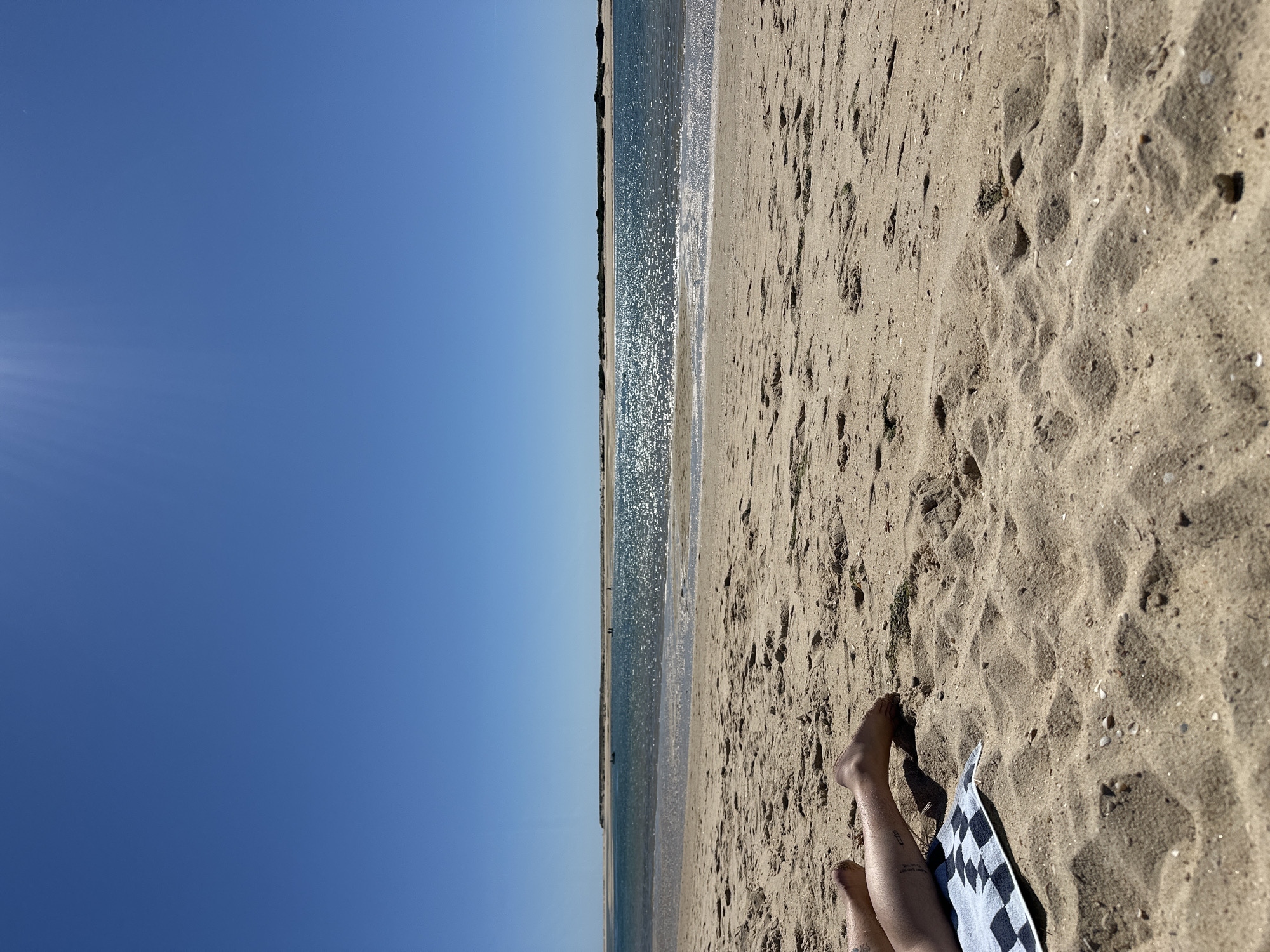 The image shows a beach scene, likely in Olhão, Portugal, indicated by the typical landscape. The perspective is from someone lying on the sand, with their legs visible in the lower right. A checkered towel is partially visible beside the legs. The sand is light brown and has numerous footprints. The ocean is in the center, with sparkling sunlight reflecting on the water. A thin strip of land or island is visible in the distance. The sky is a clear, bright blue. The image is oriented vertically.