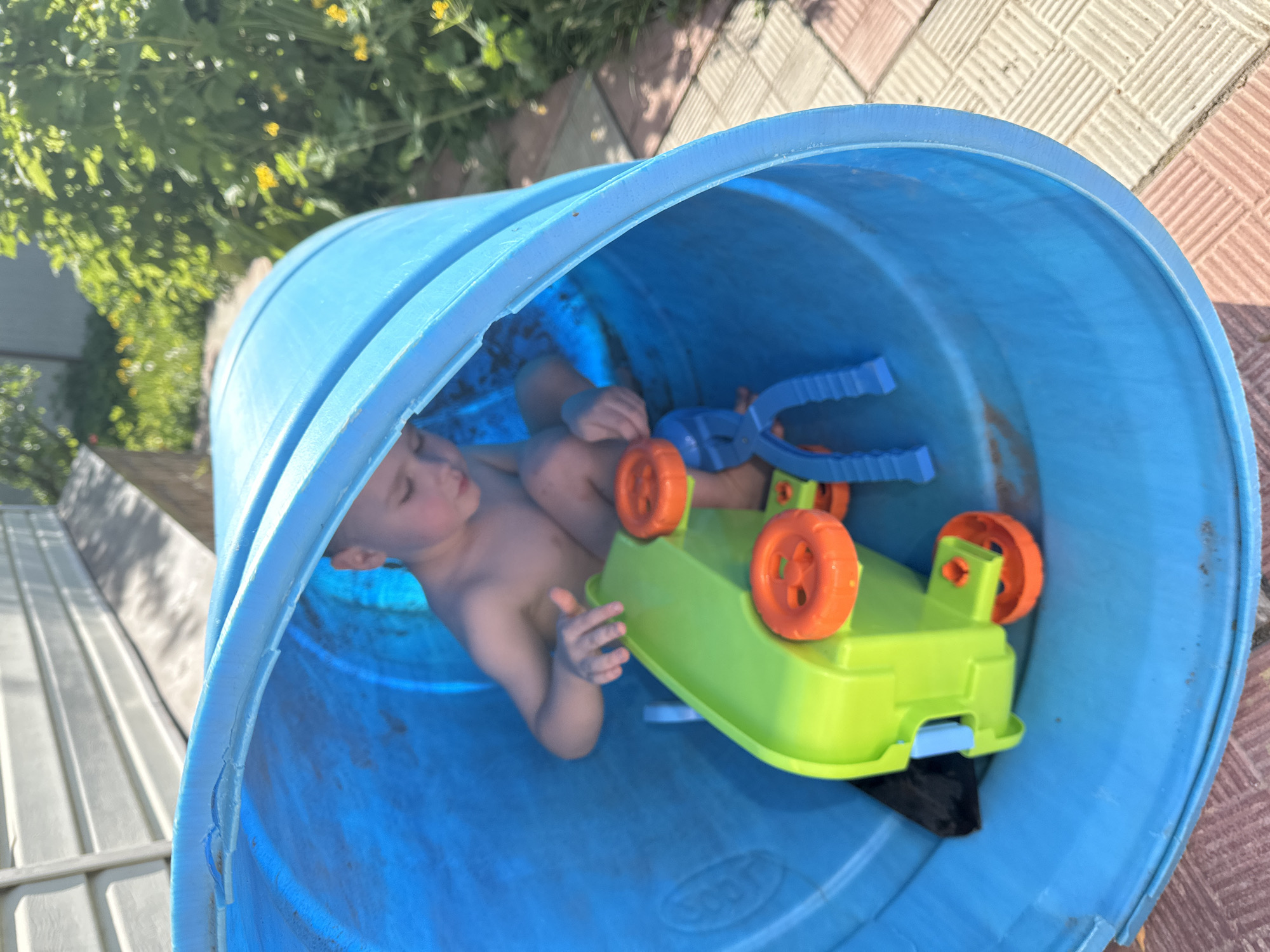 A young, light-skinned child is lying on his back inside a large, blue plastic barrel. The child is shirtless and appears to be wearing patterned blue shorts. A green plastic toy truck with orange wheels is partially on top of the child. A blue plastic toy is also nearby. The barrel is resting on a paved surface composed of light-colored blocks, near some greenery. The location of the image is unknown and there is no evidence to suggest that it is in the city of Nizhny Novgorod, Russia.