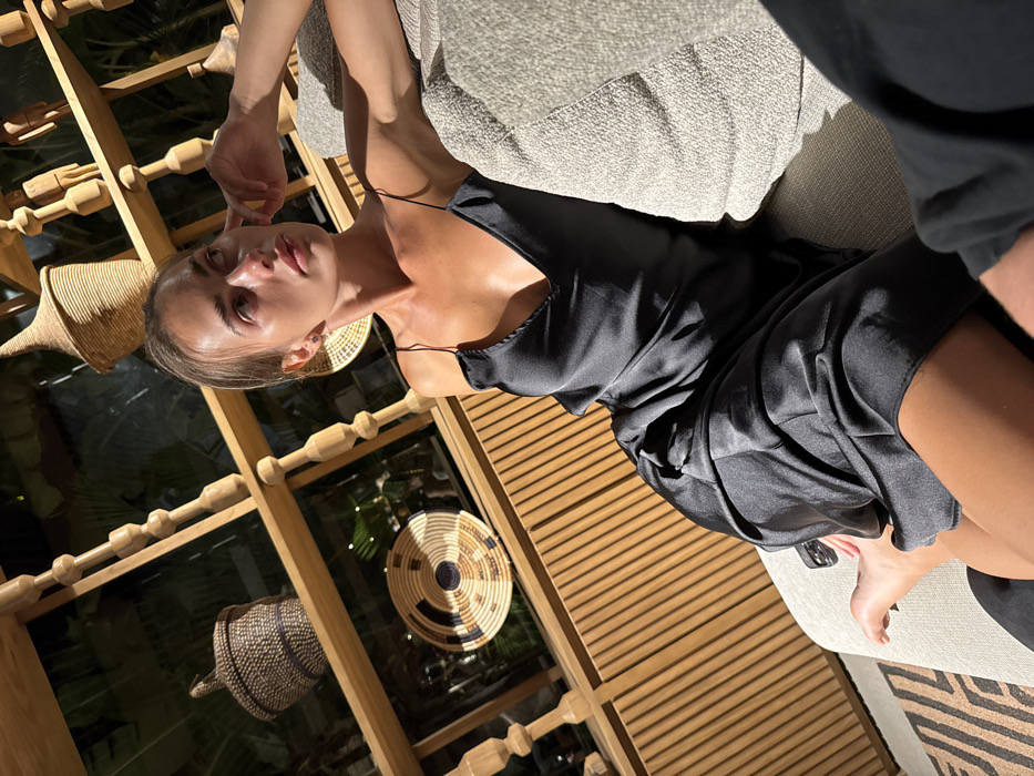 A woman in a black silky slip dress reclines on a beige couch. Her fair skin is highlighted by the light. She rests her hand near her face, gazing at the camera. Behind her is a light wood structure with decorative baskets hanging, and a window showing greenery. The couch has a textured fabric, and part of another person's arm is visible.