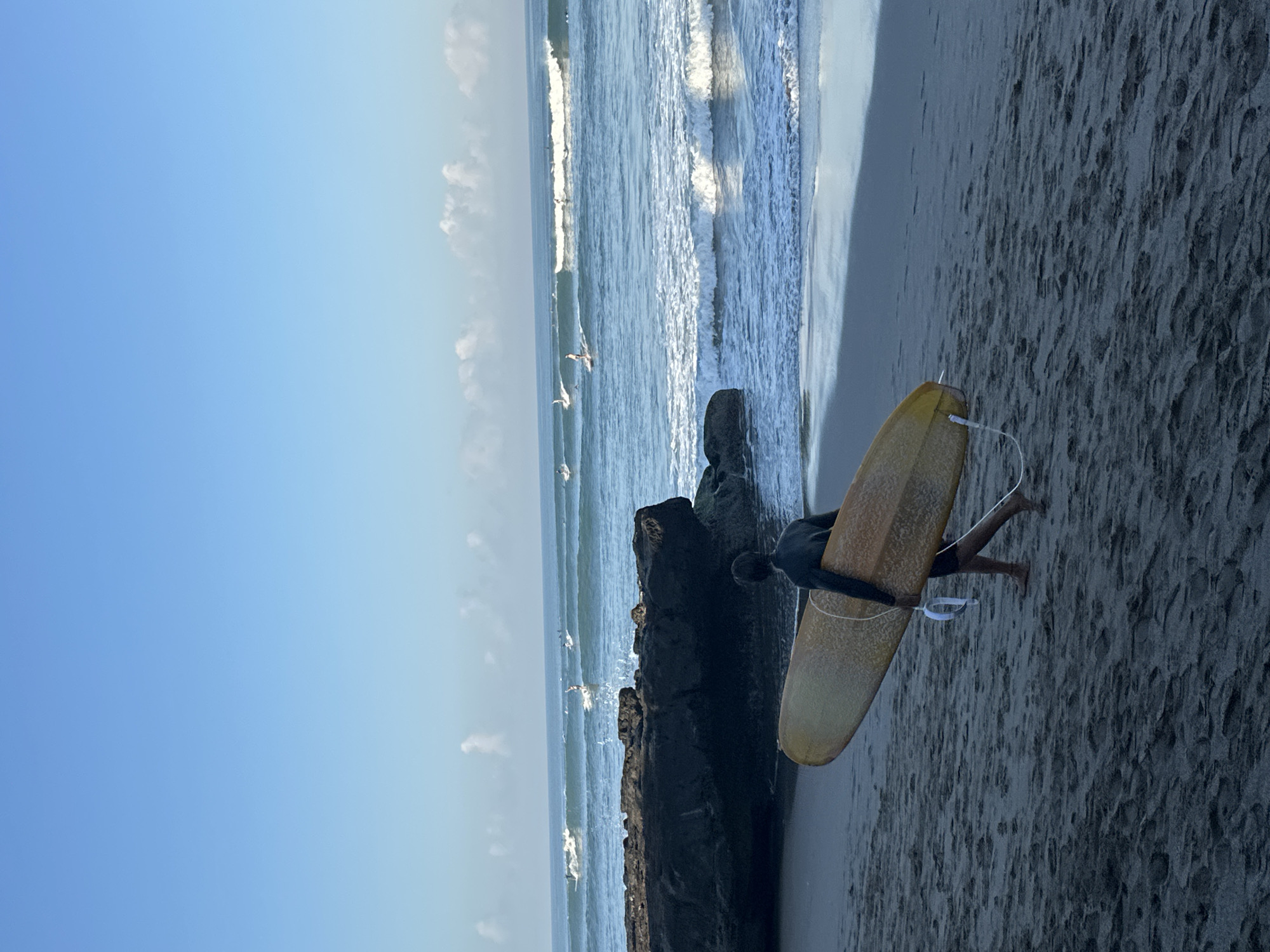 A lone surfer, wearing a black wetsuit, walks across the sandy shore of North Kuta, Indonesia, carrying a yellow surfboard. The sun casts a long shadow from the surfer onto the dark sand. The ocean is calm with small waves breaking near the shore. Several other surfers are visible in the distance, riding larger waves. A large dark rock formation juts out from the water on the left side of the image, creating a barrier between the beach and the open sea. The sky is clear and blue with a few wispy clouds.